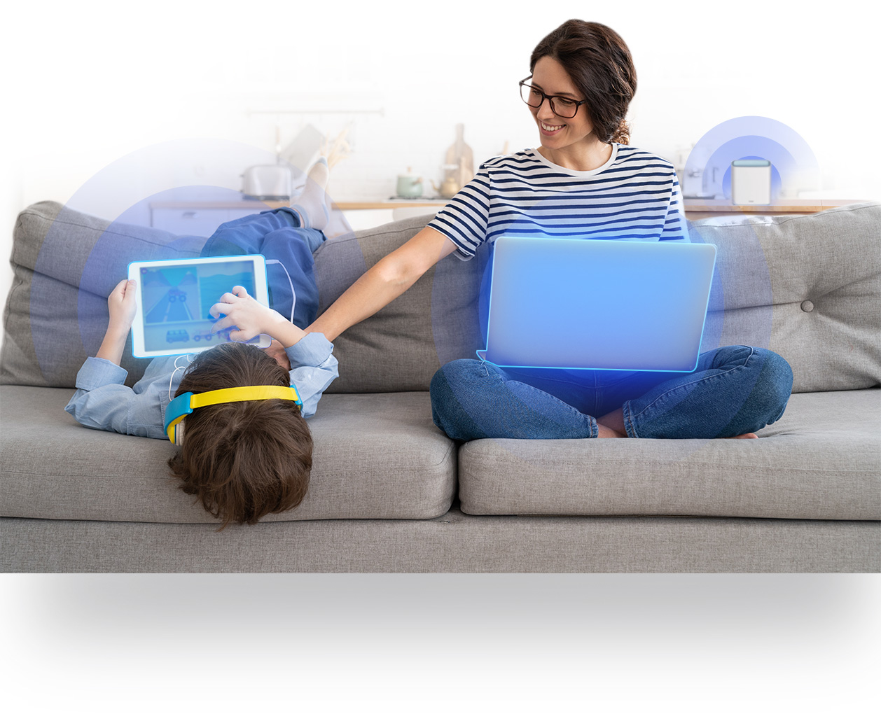 Kobieta w okularach i dziecko siedzą na kanapie. Kobieta używa laptopa, a dziecko tabletu ze słuchawkami.