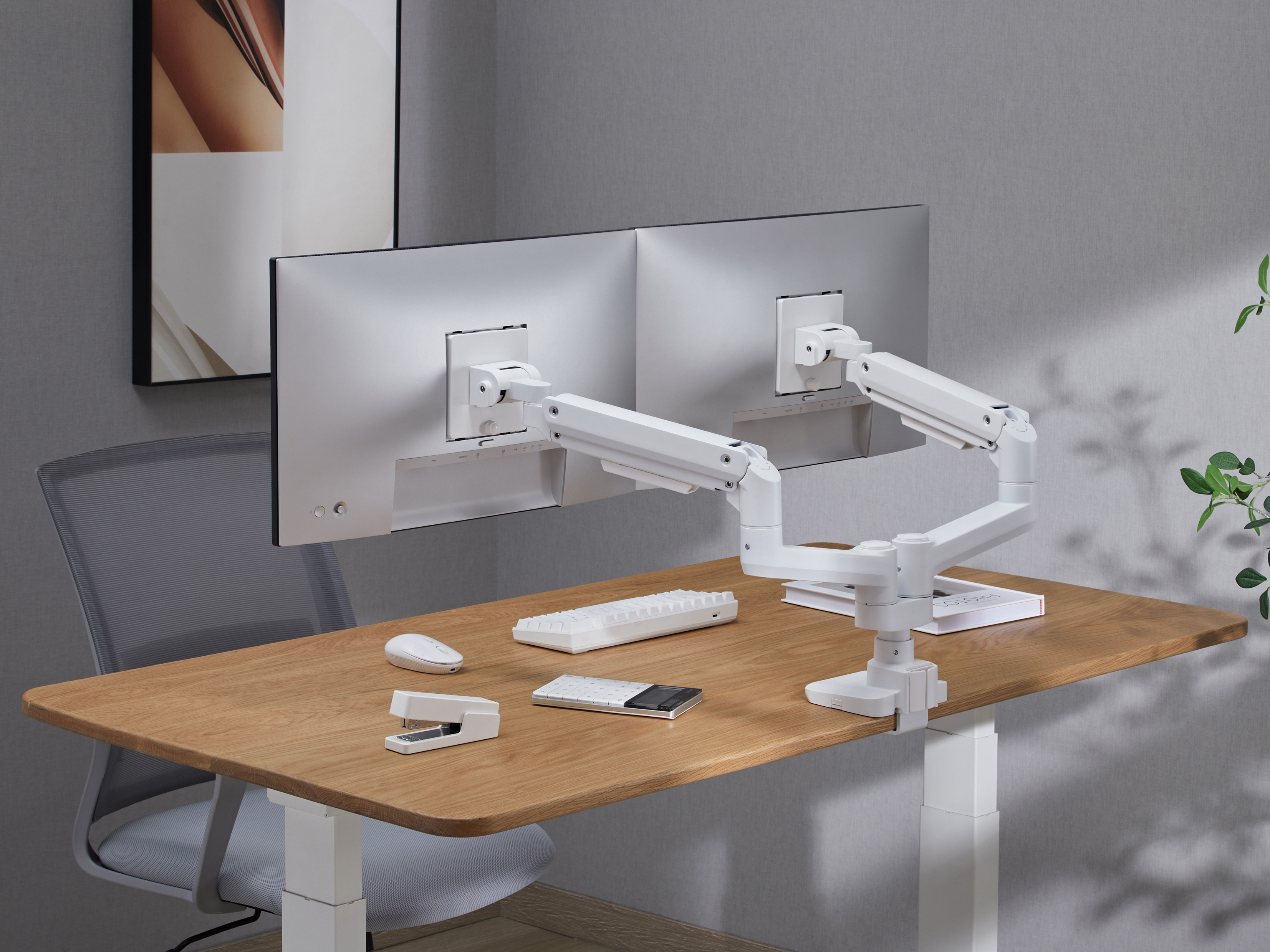 Een bureau met twee monitoren en een witte monitorarm. Er ligt een toetsenbord, muis, nietmachine en rekenmachine op het bureau.