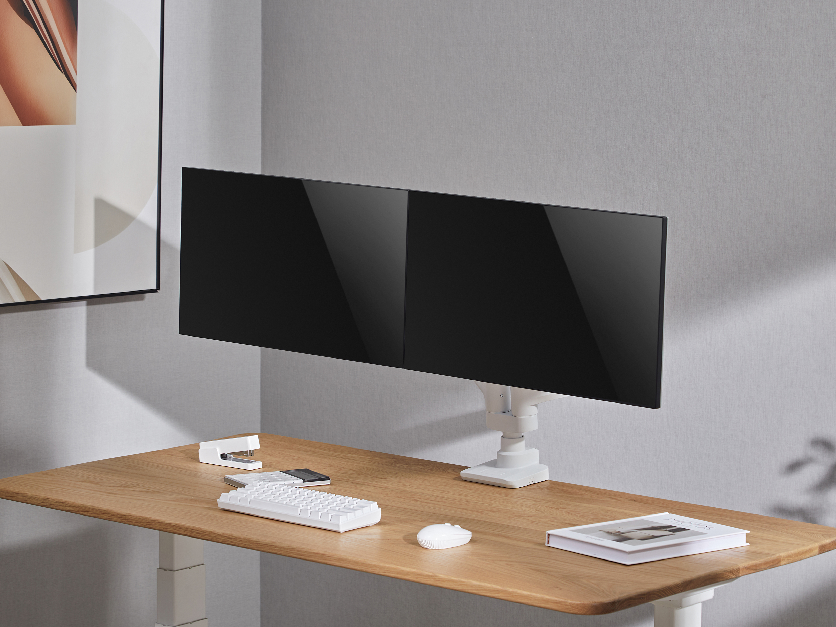 Een bureau met twee zwarte monitoren, een toetsenbord, een nietmachine en een muis. Er is een boek en een kunstwerk aanwezig.