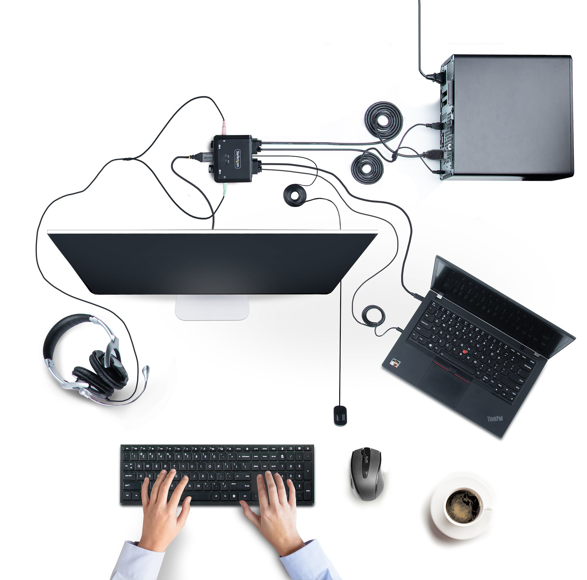Draufsicht auf einen Arbeitsplatz mit Monitor, Tastatur, Laptop, Kaffeetasse, Kopfhörern und Computer mit Kabeln.