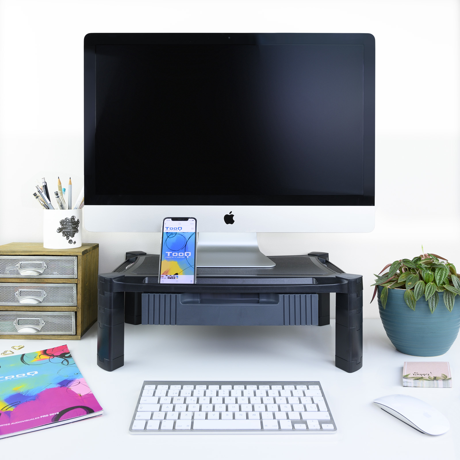 Configuración de escritorio con monitor, teléfono, teclado y planta. El teléfono muestra una app. El escritorio es blanco.