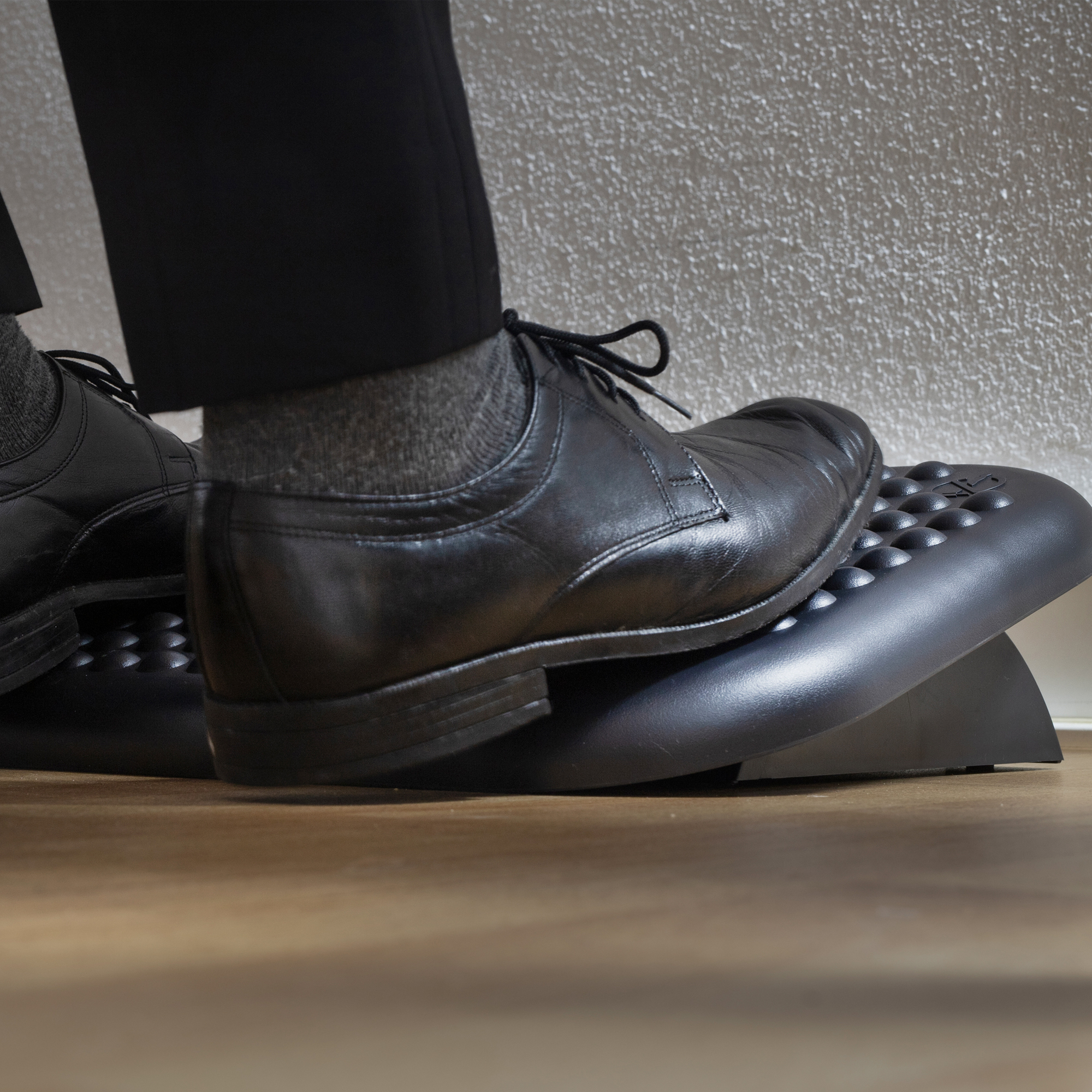 Pies de una persona con zapatos negros en un reposapiés. El fondo es una pared blanca y un suelo marrón.