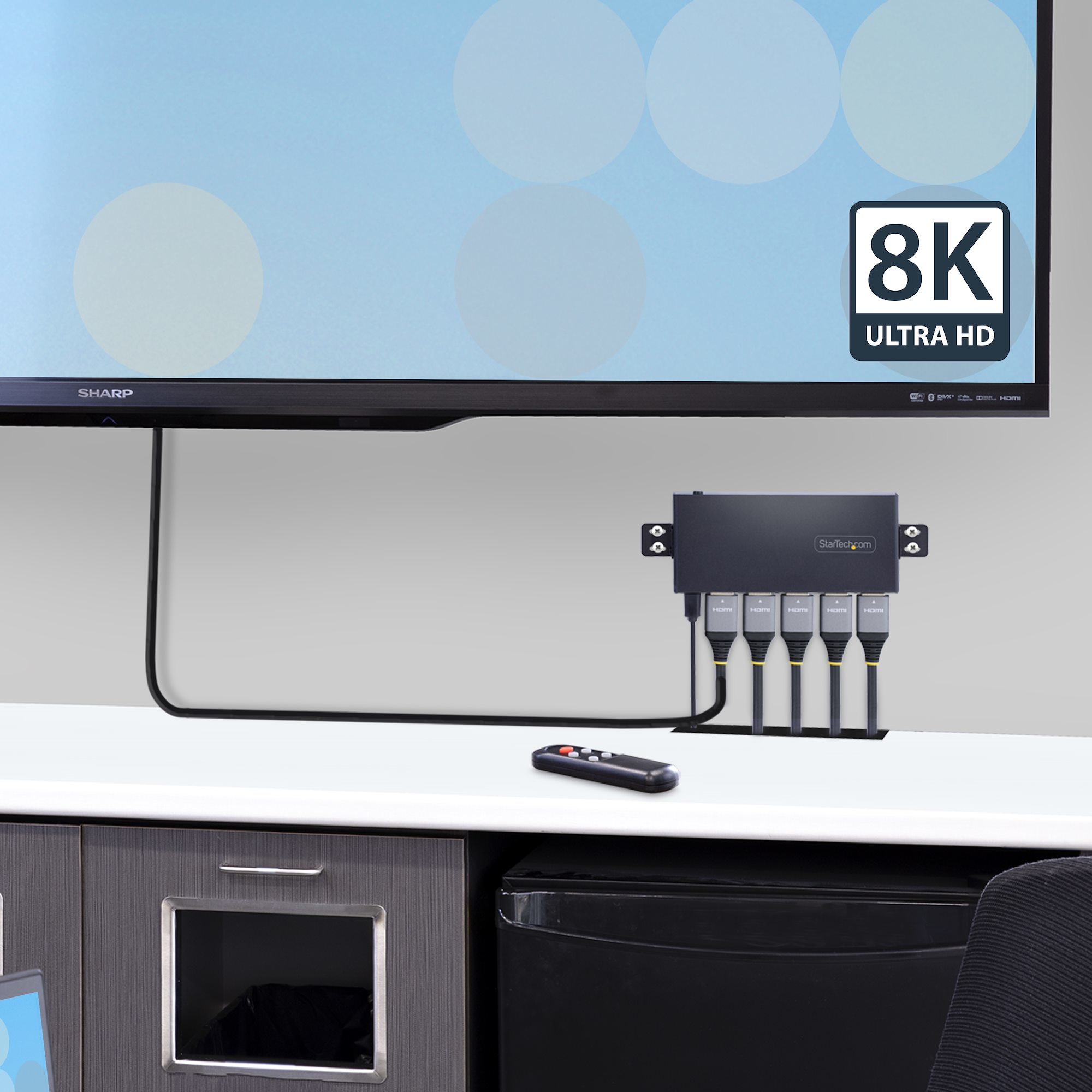 Una TV 8K Ultra HD con uno splitter HDMI nero, telecomando e cavi su una scrivania bianca.