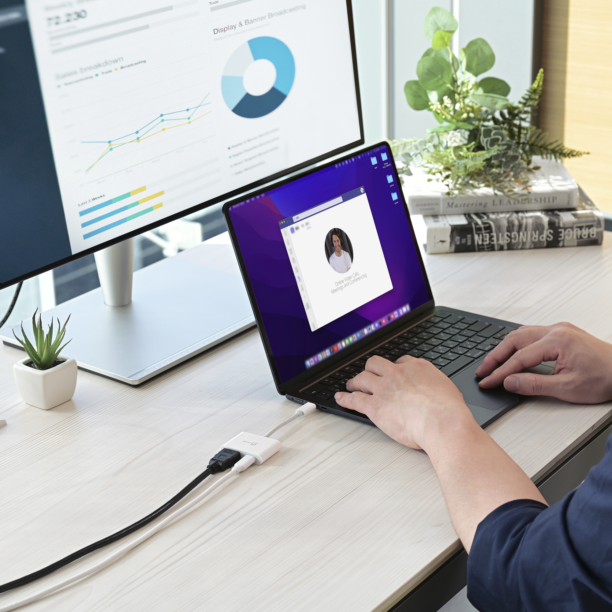 Laptop z rozmową wideo, podłączony do monitora z wykresami. Osoba korzysta z laptopa. Rośliny i książki w tle.