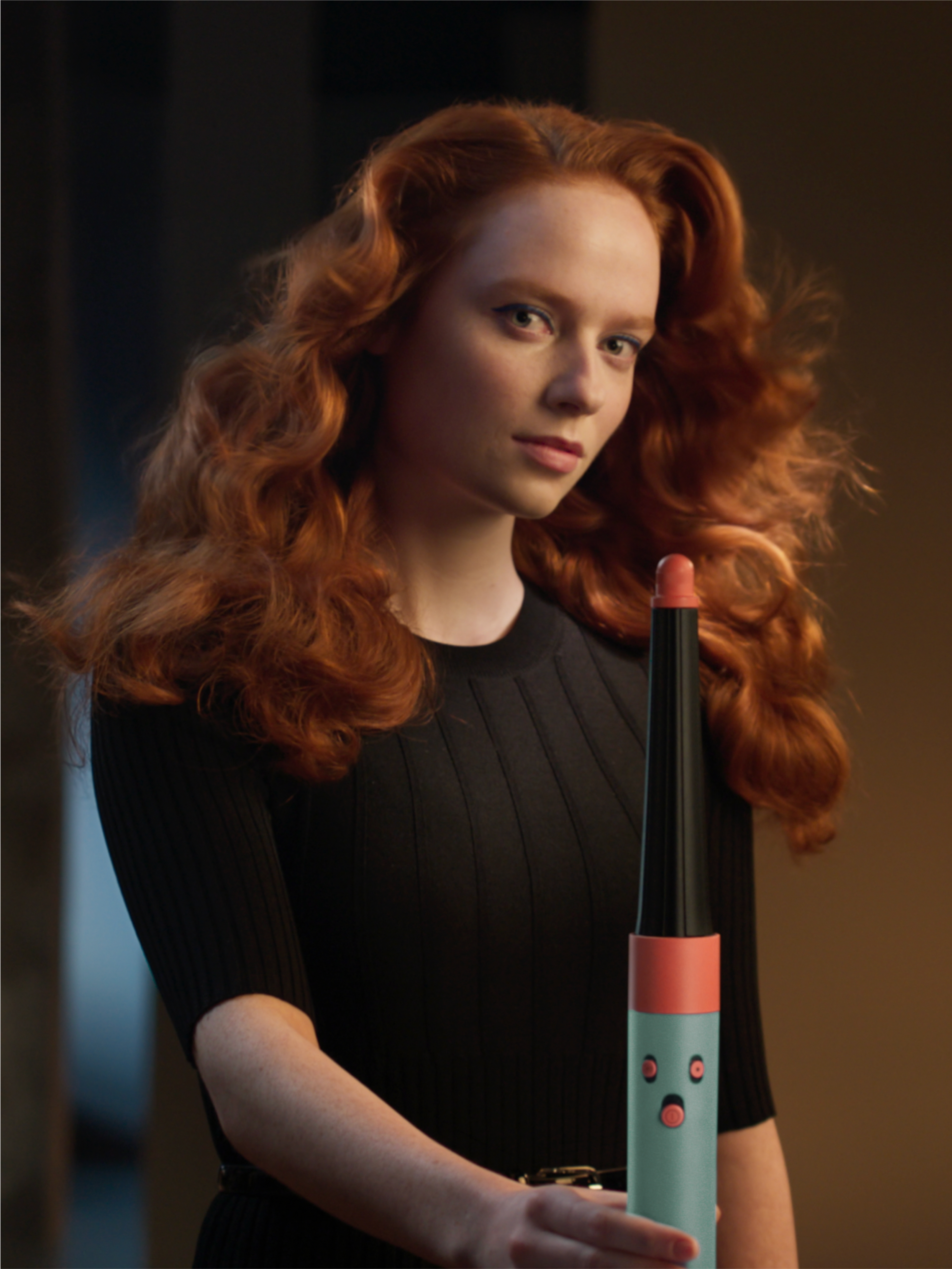 Kobieta z rudymi włosami trzyma narzędzie do stylizacji. Ma na sobie czarny top. Tło jest rozmyte brązowe.