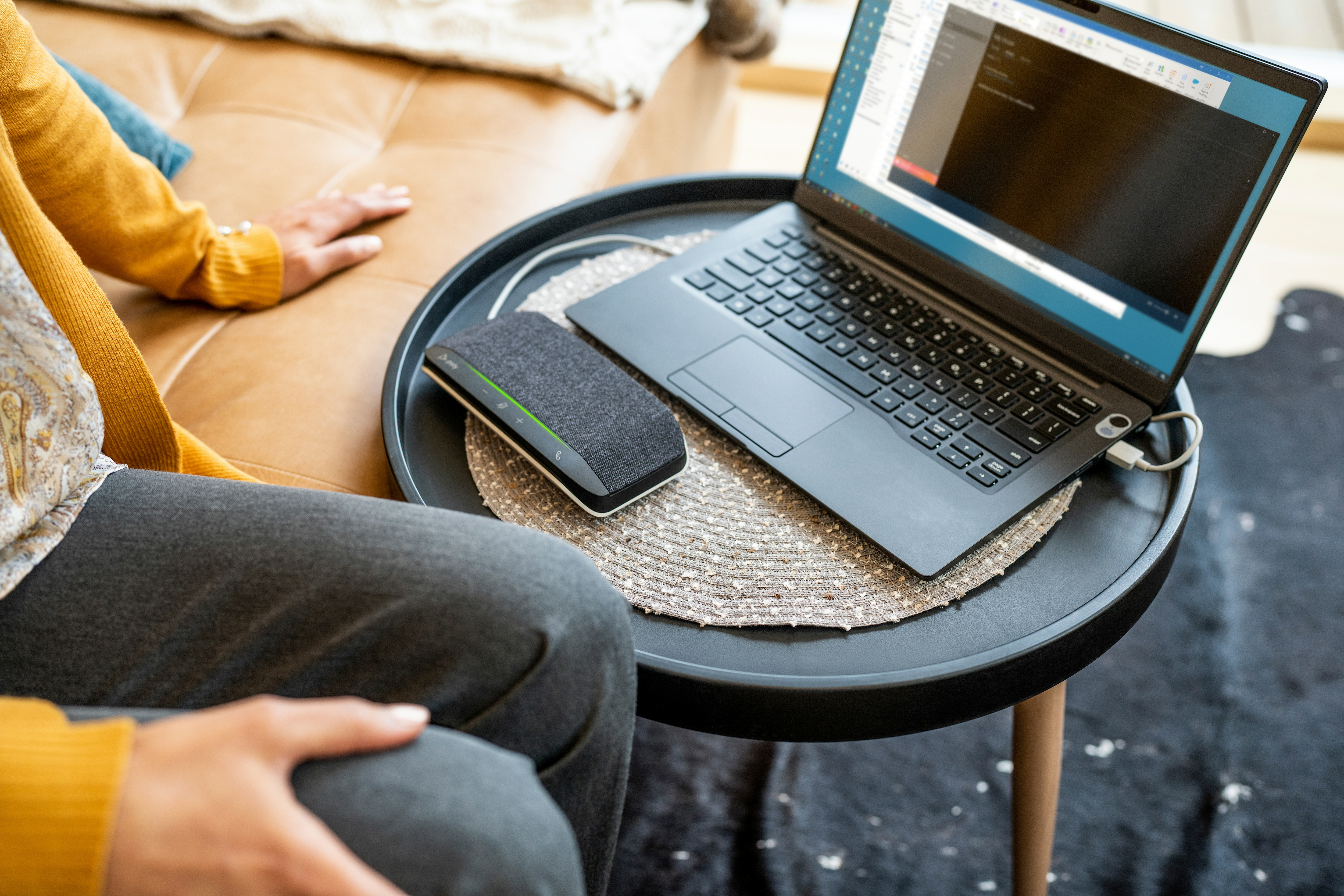 Un laptop e un altoparlante su un tavolino nero. Una persona è seduta nelle vicinanze. Lo sfondo è un divano e un tappeto.
