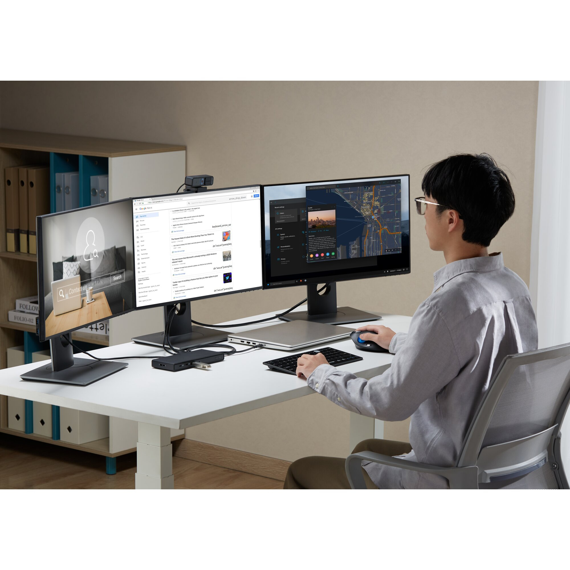 Ein Mann sitzt an einem Schreibtisch mit zwei Monitoren und benutzt eine Tastatur und eine Maus. Die Bildschirme zeigen Websites und Karten.