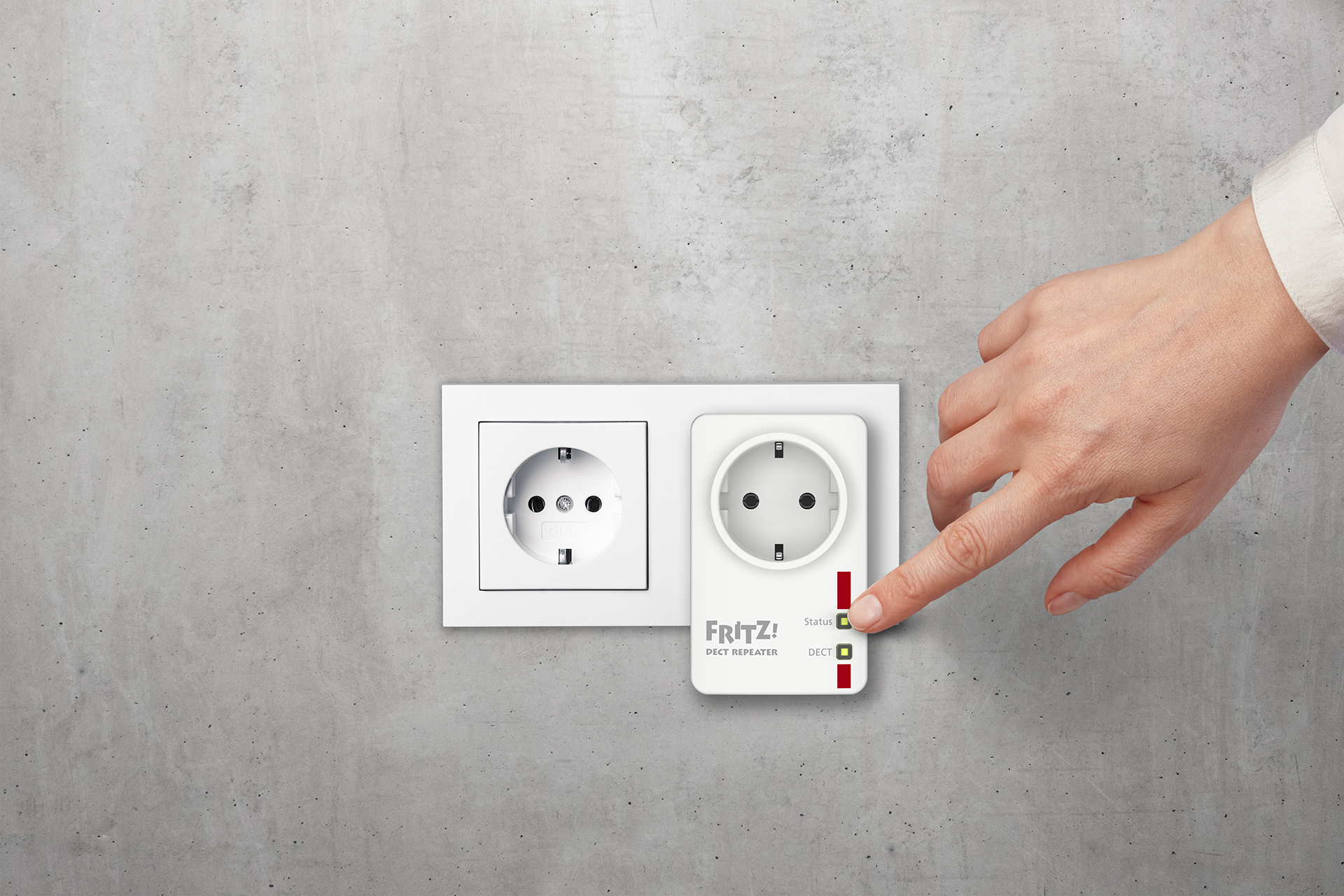 Mano señalando un repetidor FRITZ! DECT blanco conectado a un enchufe de pared. Fondo de hormigón.