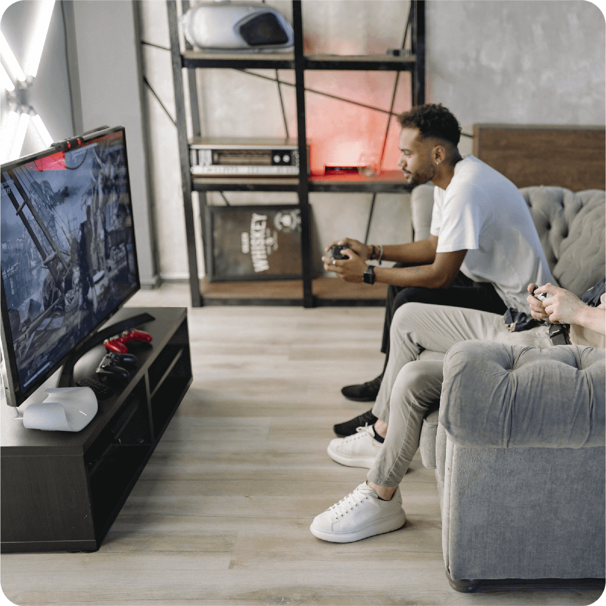 Dwóch mężczyzn na kanapie gra w gry wideo. Telewizor wyświetla grę. Za nimi półki z przedmiotami.