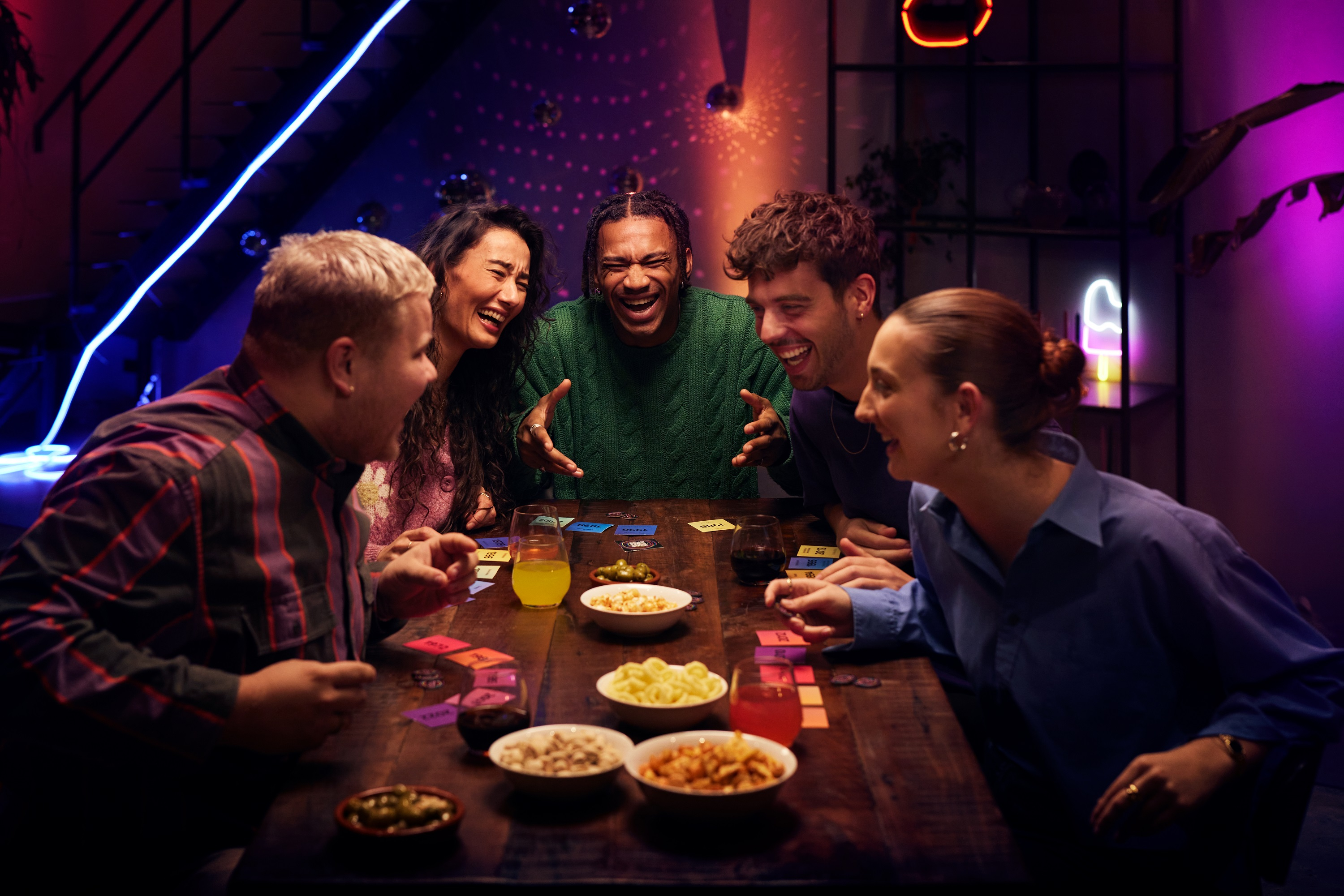 Grupa przyjaciół śmiejąca się wokół stołu, grająca w grę. Na stole przekąski i napoje. Słabo oświetlony pokój.