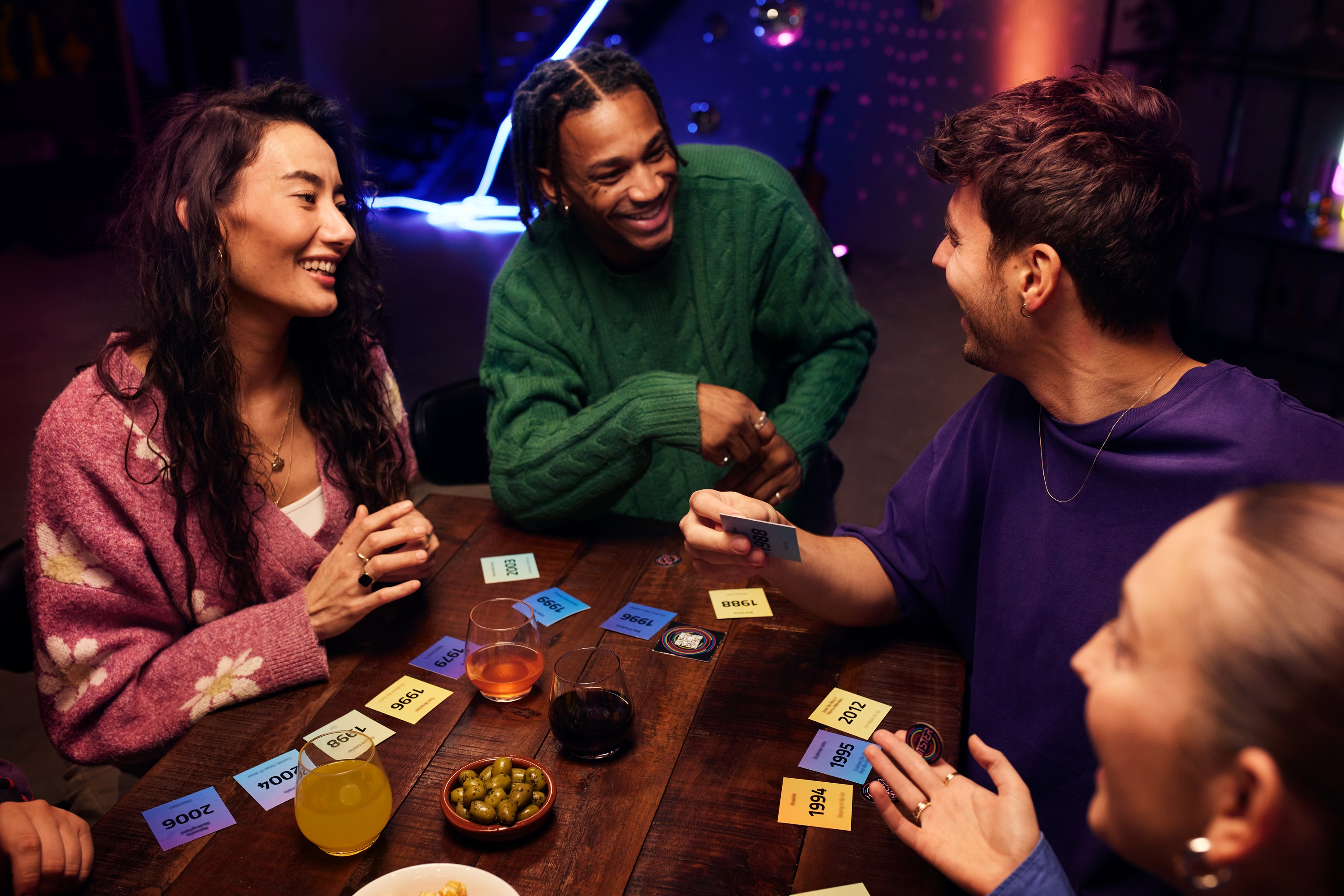 Cztery osoby grają w grę planszową. Na stole leżą karty z latami. Uśmiechają się i wchodzą w interakcje.