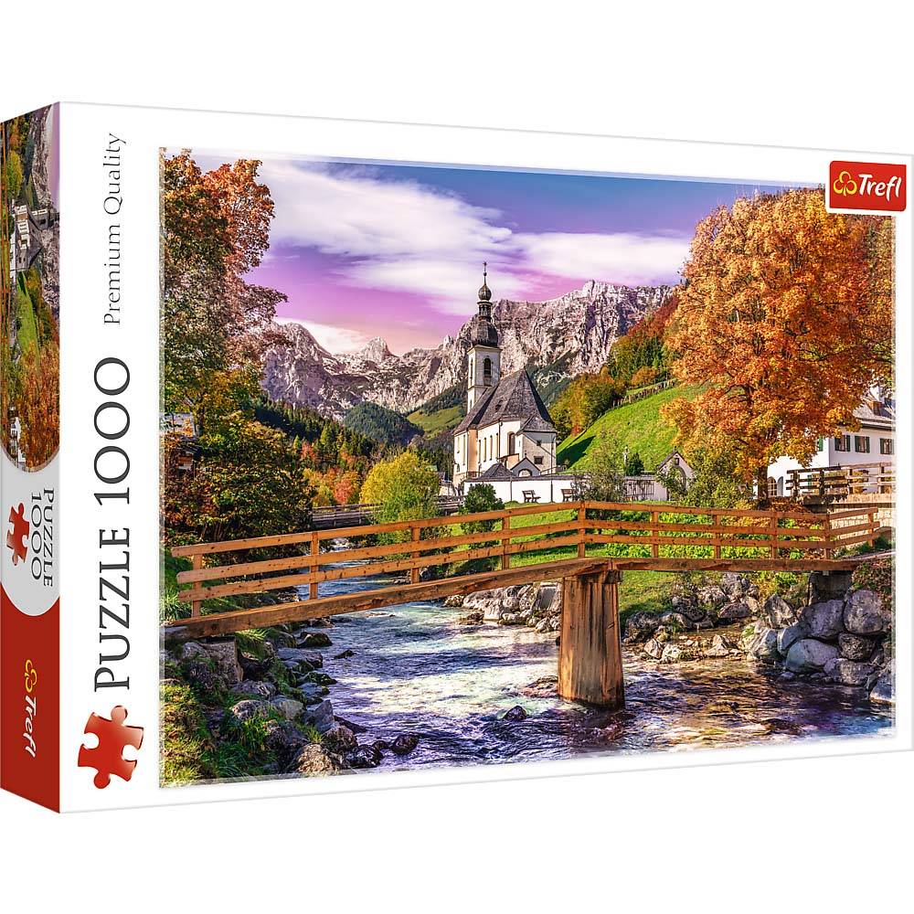 Scatola di puzzle mostra chiesa vicino alle montagne. Un ponte di legno attraversa un fiume. Alberi autunnali incorniciano la scena. Cielo vivido.