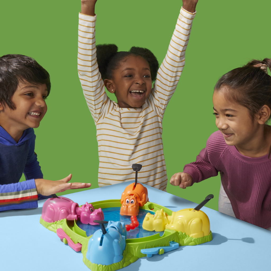 Tre bambini giocano a Hungry Hungry Hippos. Una ragazza esulta con le braccia alzate. Il gioco ha ippopotami colorati e biglie.