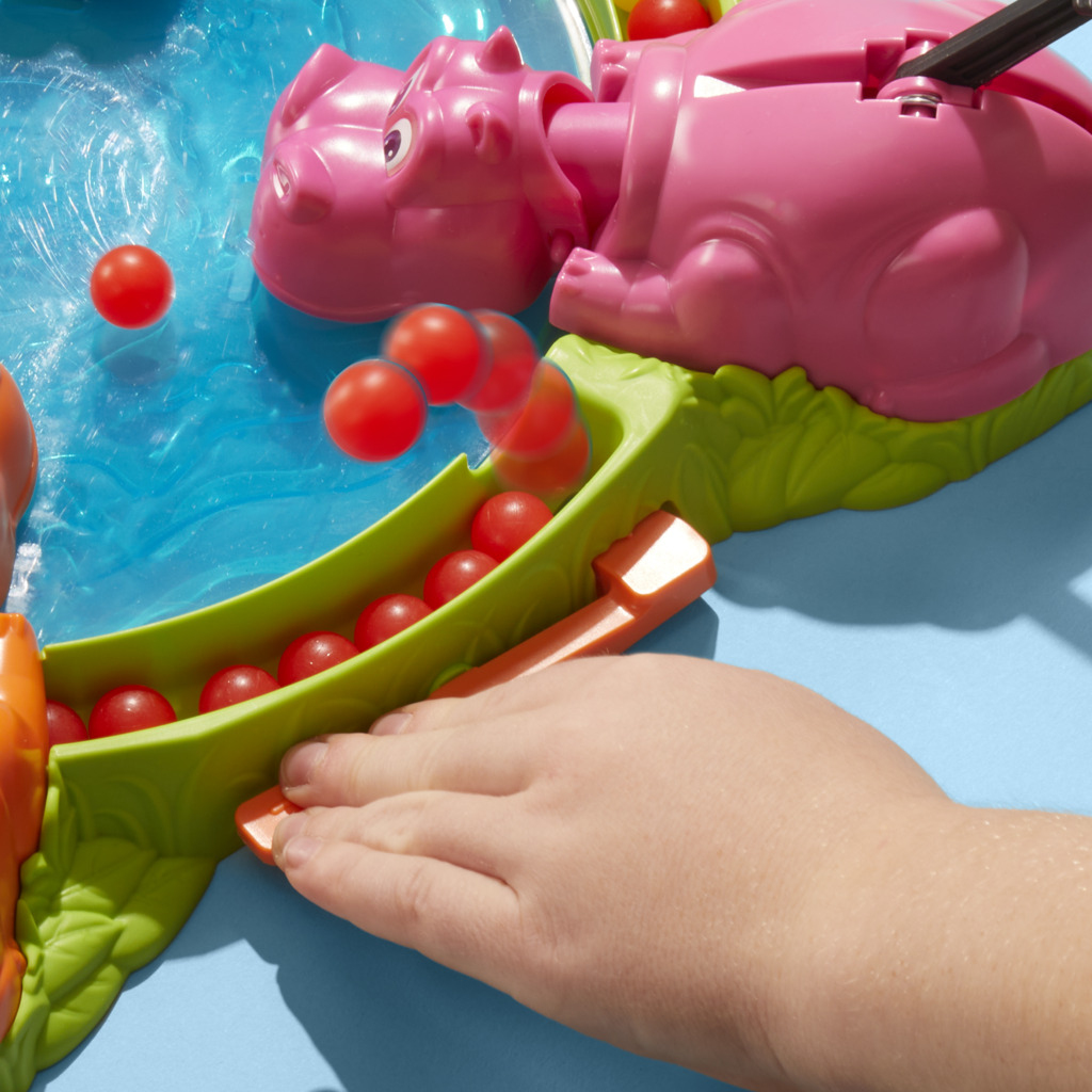 La mano di un bambino interagisce con un giocattolo a forma di ippopotamo rosa. Palline rosse sono nel vassoio verde e vengono lanciate. Sfondo blu.