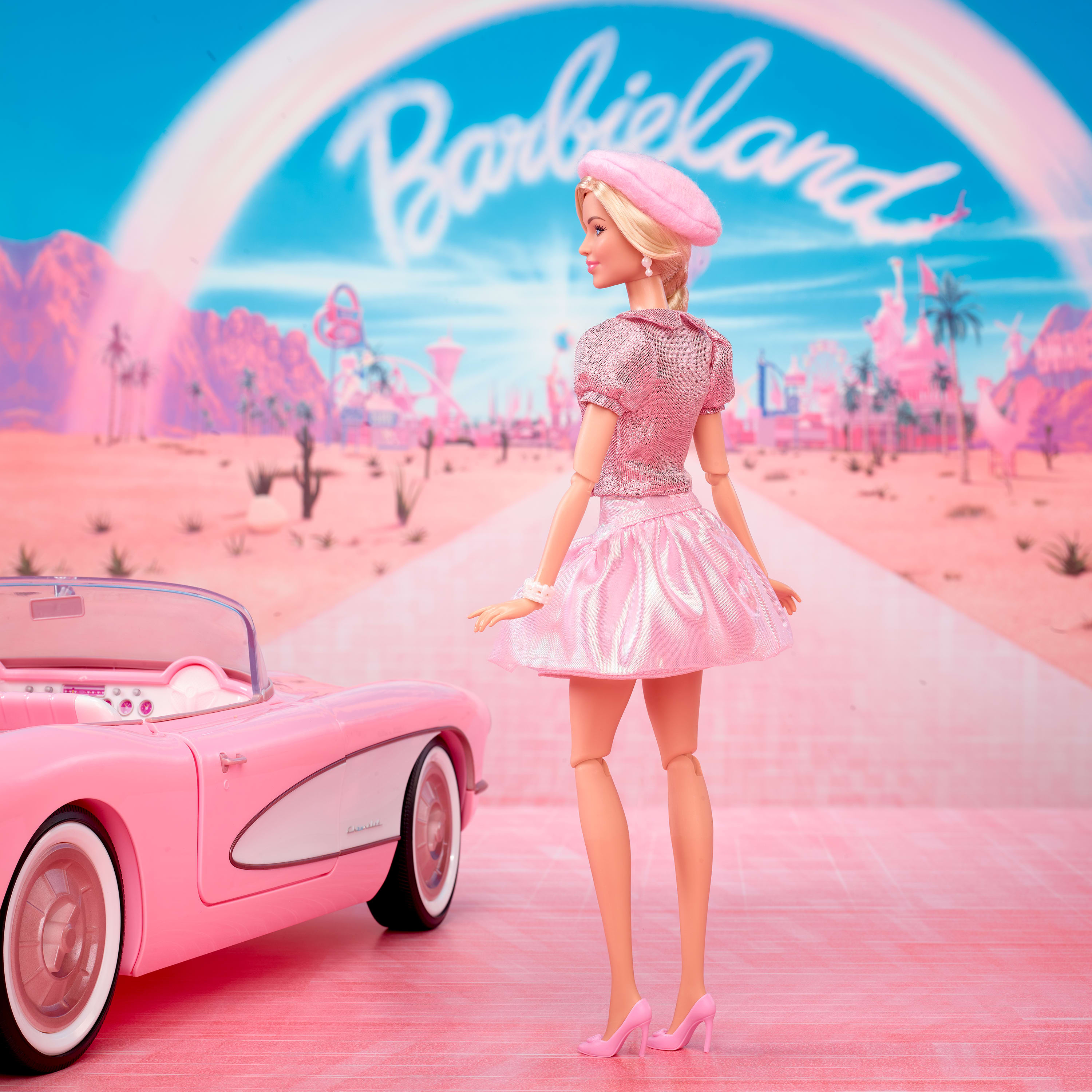 Una muñeca Barbie con atuendo rosa está junto a un coche rosa en un paisaje rosa. El cielo es azul con un arcoíris.
