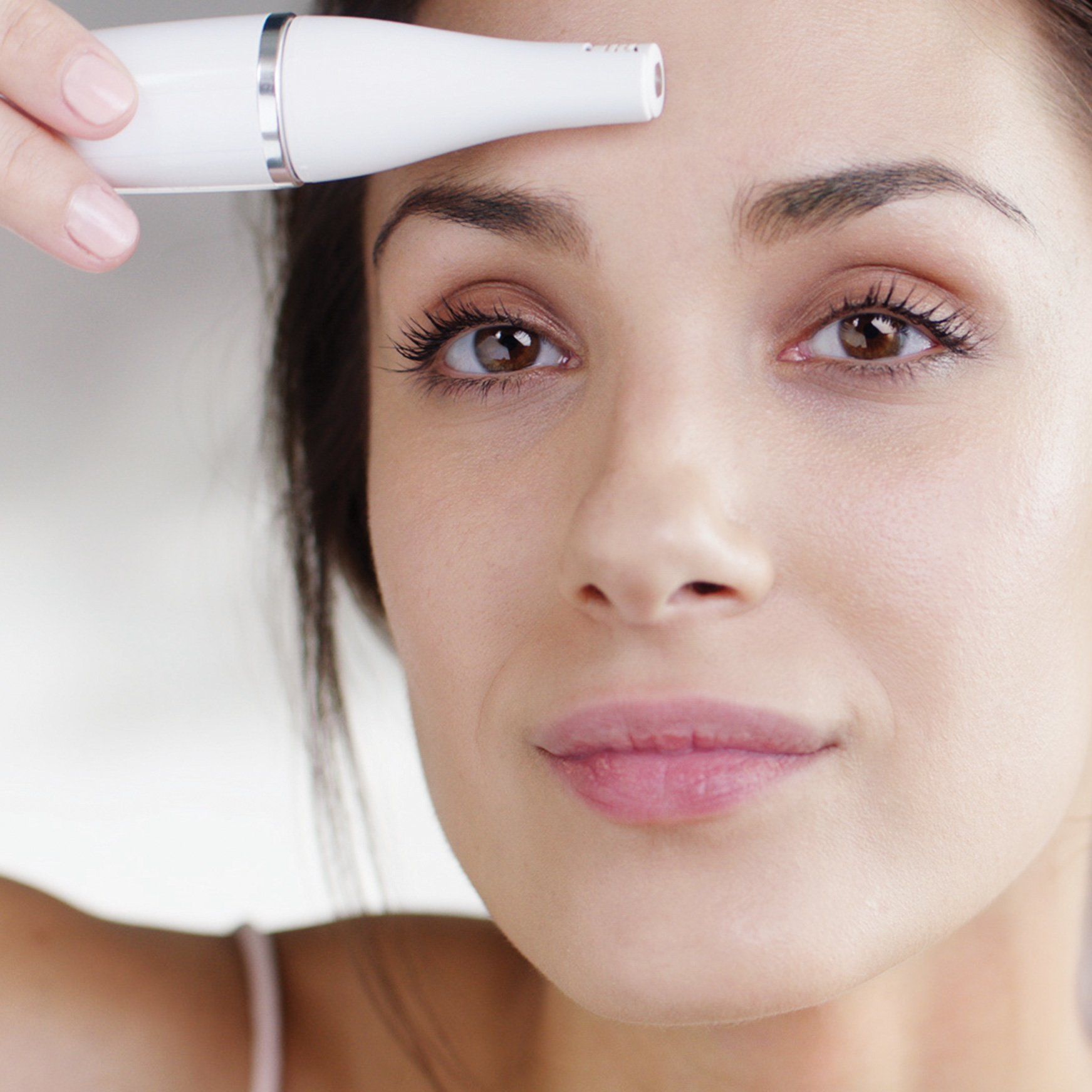 Kobieta trzyma białe urządzenie kosmetyczne blisko czoła, patrząc w kamerę. Jej twarz jest ostra, z brązowymi oczami.