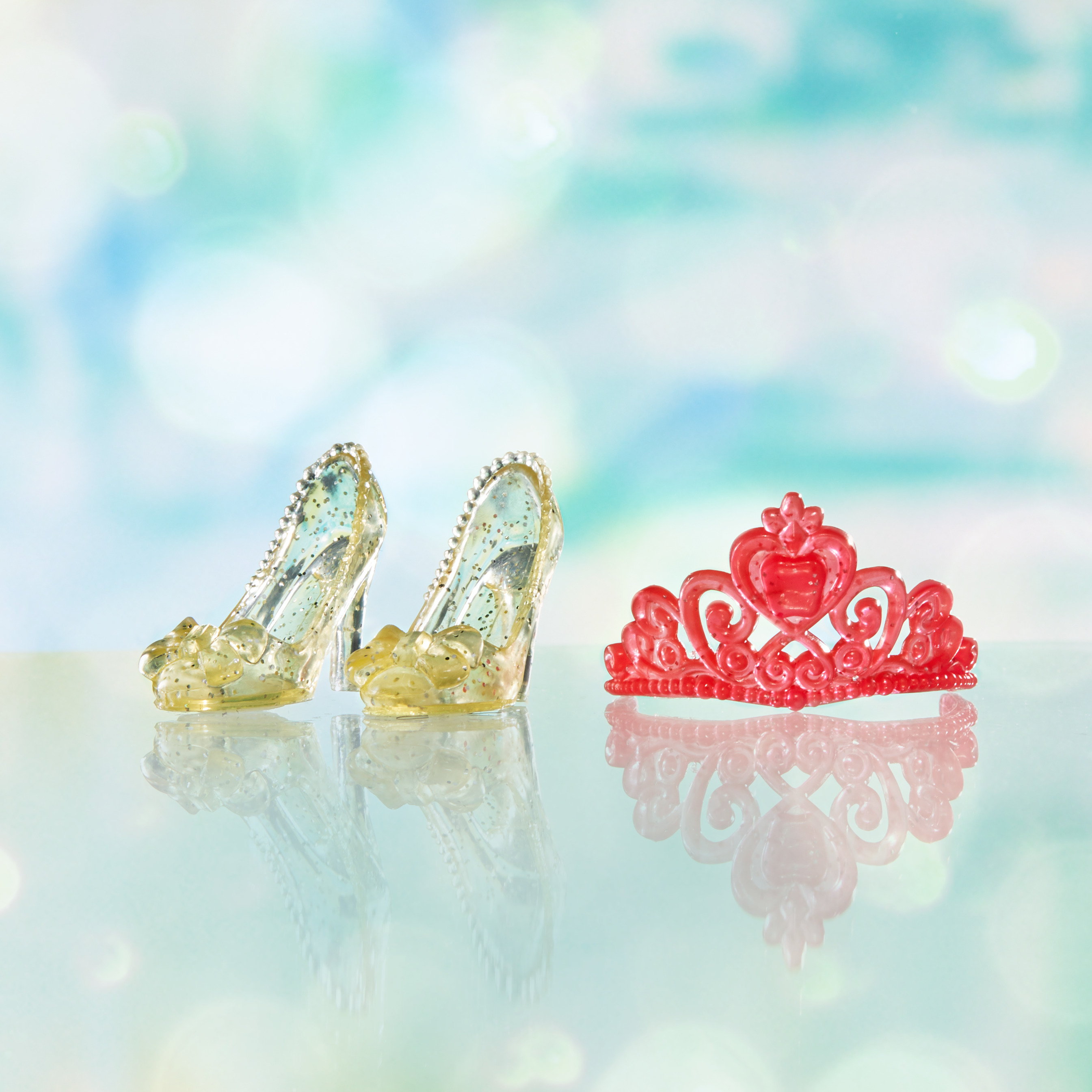 L'immagine mostra scarpette di cristallo giocattolo e una tiara rossa su una superficie riflettente, sfondo sfocato.