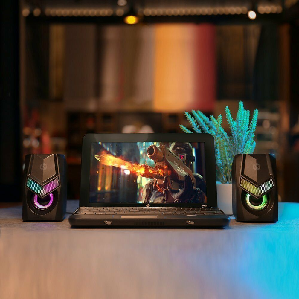 Portátil con pantalla de juego, flanqueado por altavoces con luces de colores. Una planta está detrás del portátil. El fondo está borroso.