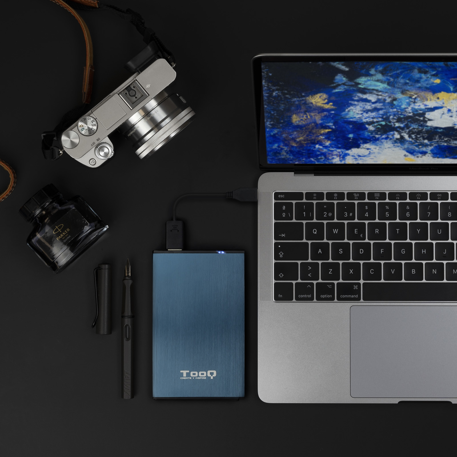 Una laptop, cámara, pluma y disco duro externo están dispuestos sobre una superficie negra.