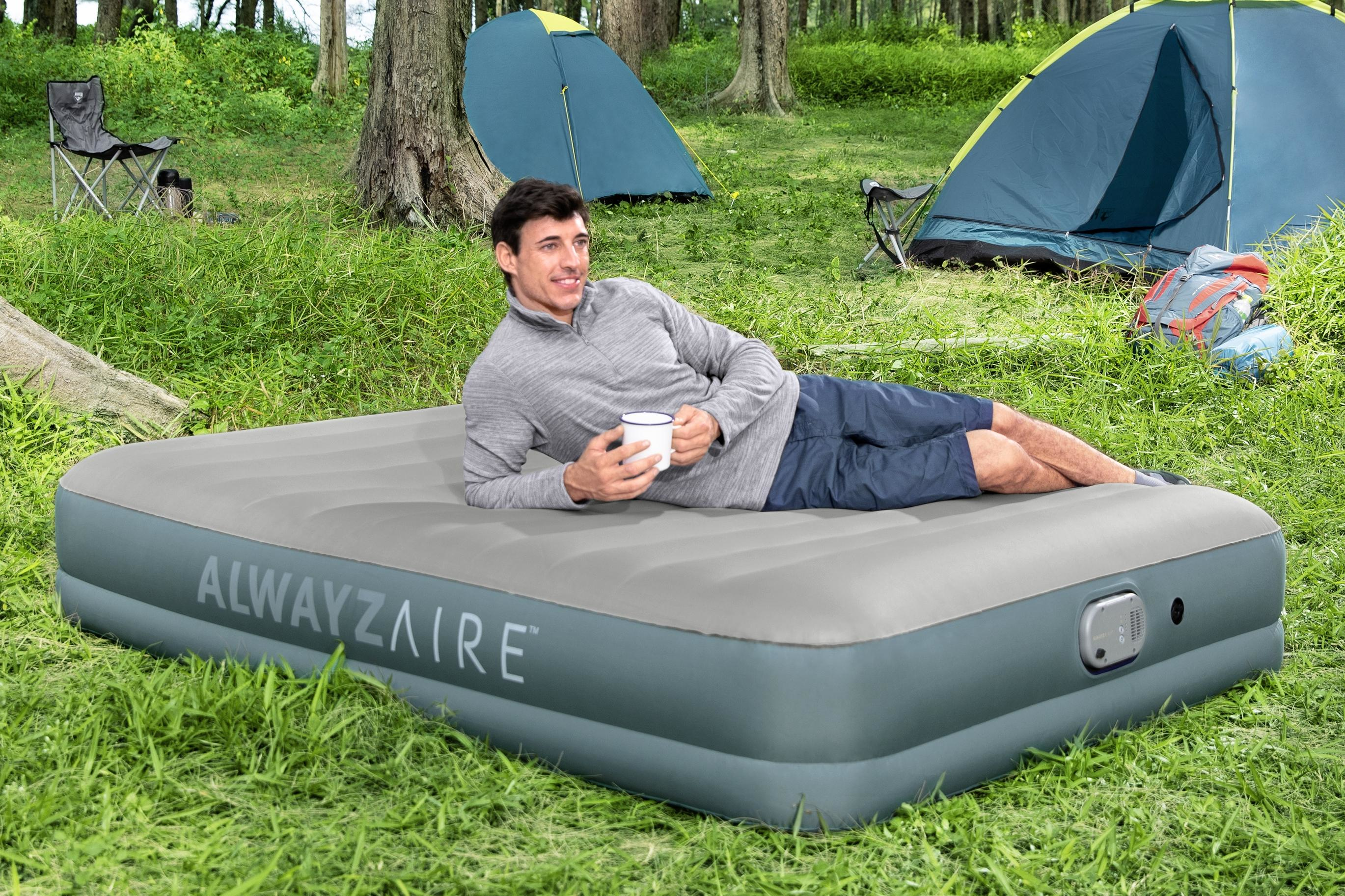 Een man ligt op een luchtmatras in een bos en houdt een mok vast. Tenten en campingstoelen op de achtergrond.