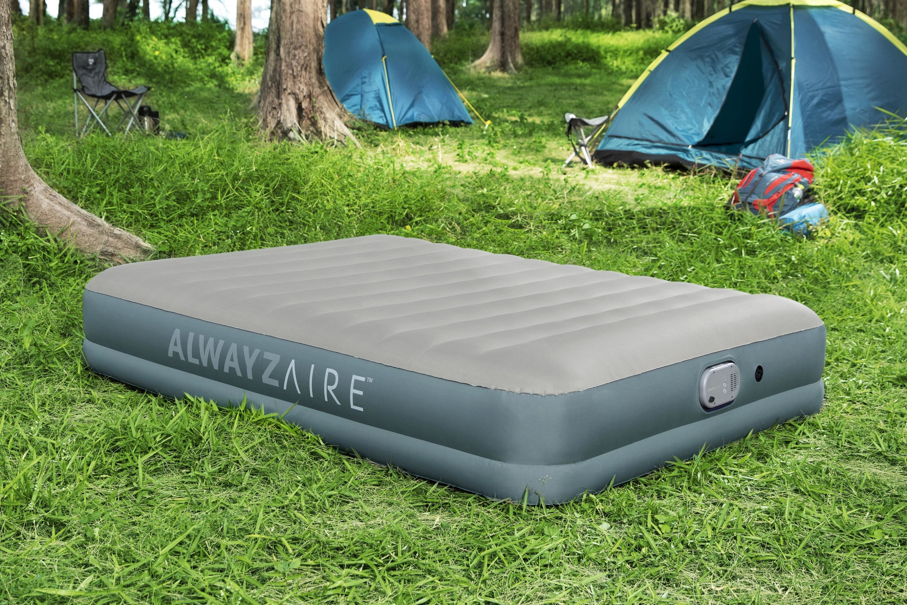 Een luchtmatras op groen gras in een bos. Tenten en campingstoelen staan op de achtergrond.