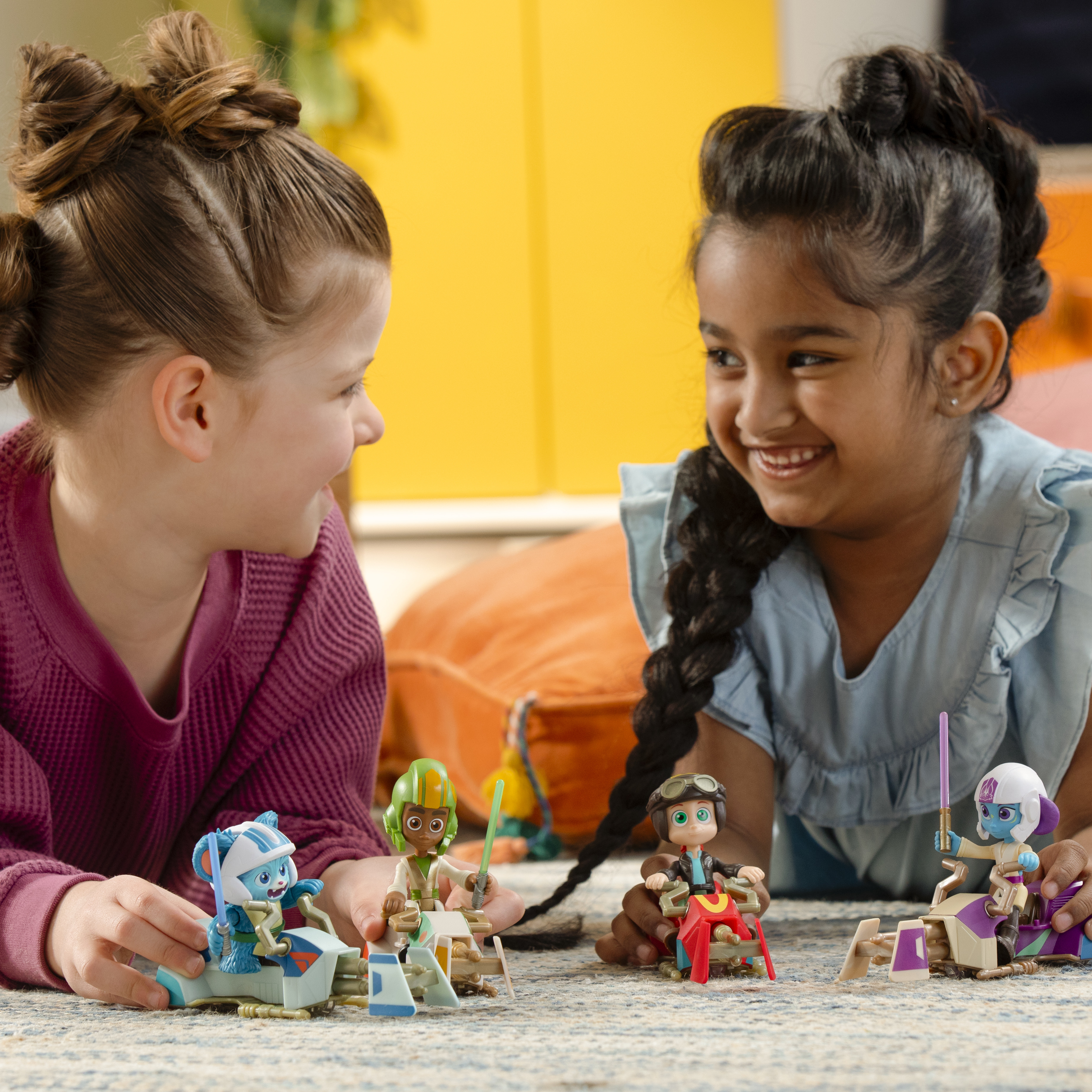 Dwie uśmiechnięte dziewczynki bawią się figurkami na dywanie. Figurki jadą pojazdami, z mieczami świetlnymi, na tle żółtego.