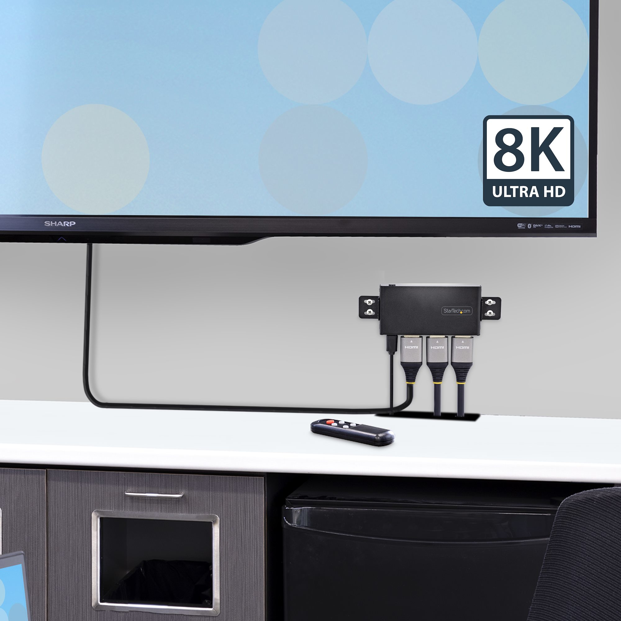 Un televisor 8K Ultra HD con un concentrador HDMI conectado y un control remoto en un escritorio blanco, con un armario debajo.