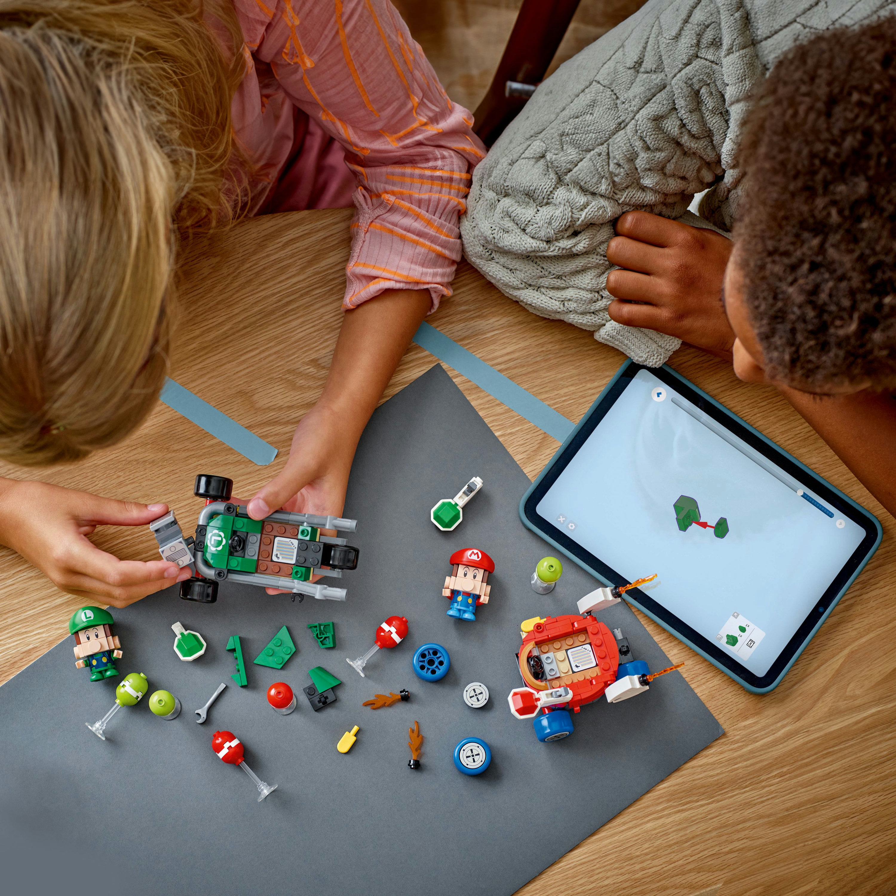 Dwoje dzieci składa zestawy Lego Mario na drewnianym stole. Tablet wyświetla instrukcje. Klocki Lego są rozrzucone.