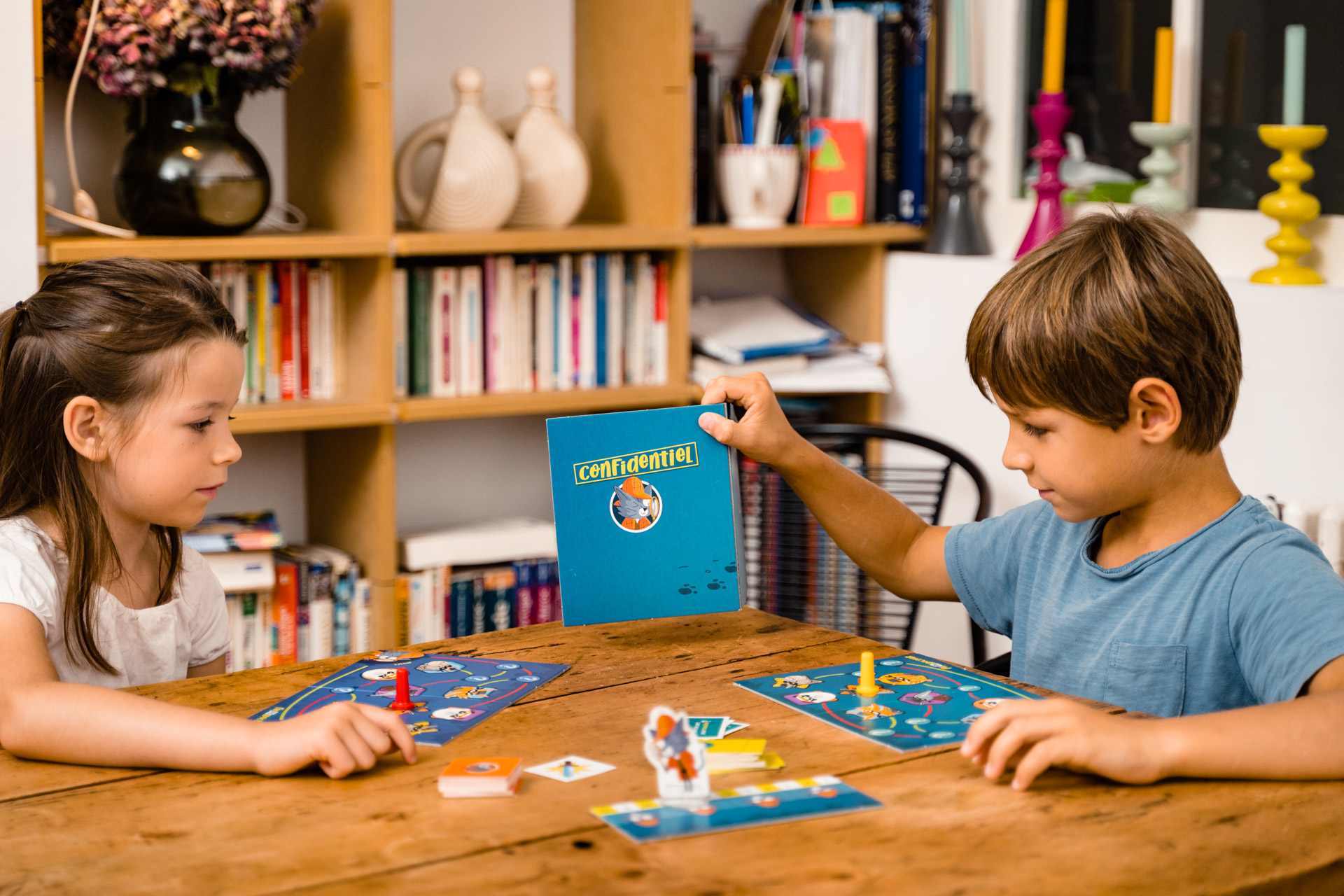 Zwei Kinder spielen ein Brettspiel an einem Holztisch. Eines hält ein blaues Buch mit der Aufschrift 'Confidential'.