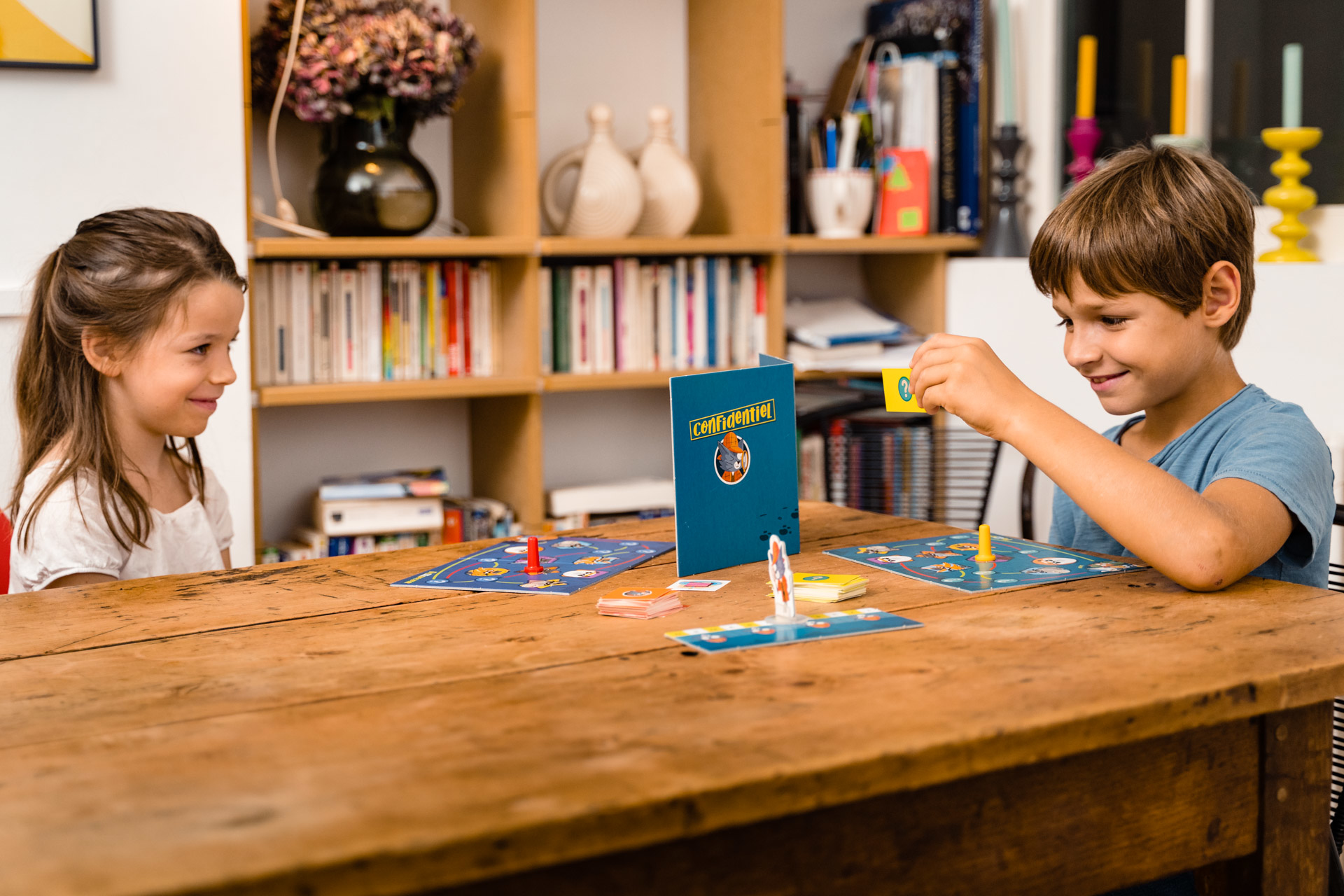 Zwei Kinder spielen ein Brettspiel. Mädchen lächelt Jungen an, der eine Karte hält. Spielbrett und Buch auf Holztisch.