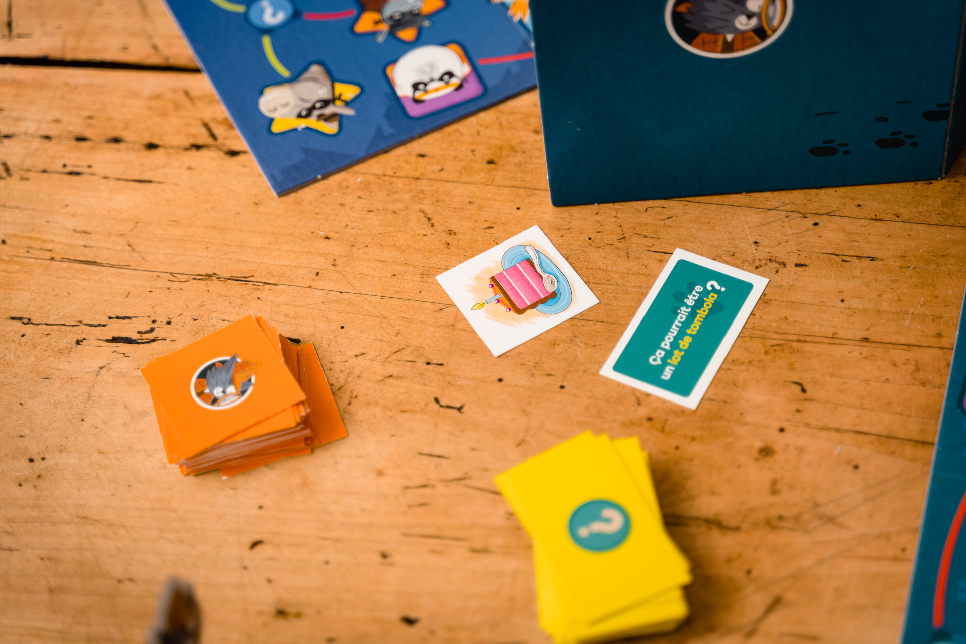 Holztisch mit Spielsteinen: Karten, Kacheln mit Illustrationen und eine blaue Spielbox. Karten sind orange, gelb und weiß.