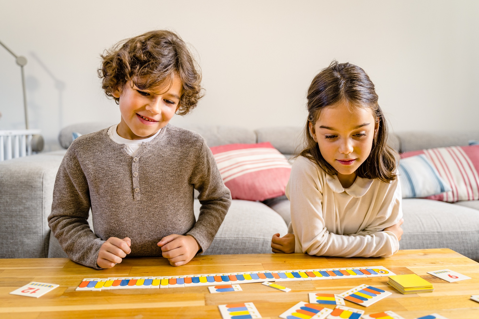 Due bambini giocano a un gioco da tavolo su un tavolo di legno. Un ragazzo con i capelli ricci sorride. Una ragazza guarda il gioco. Un divano è sullo sfondo.
