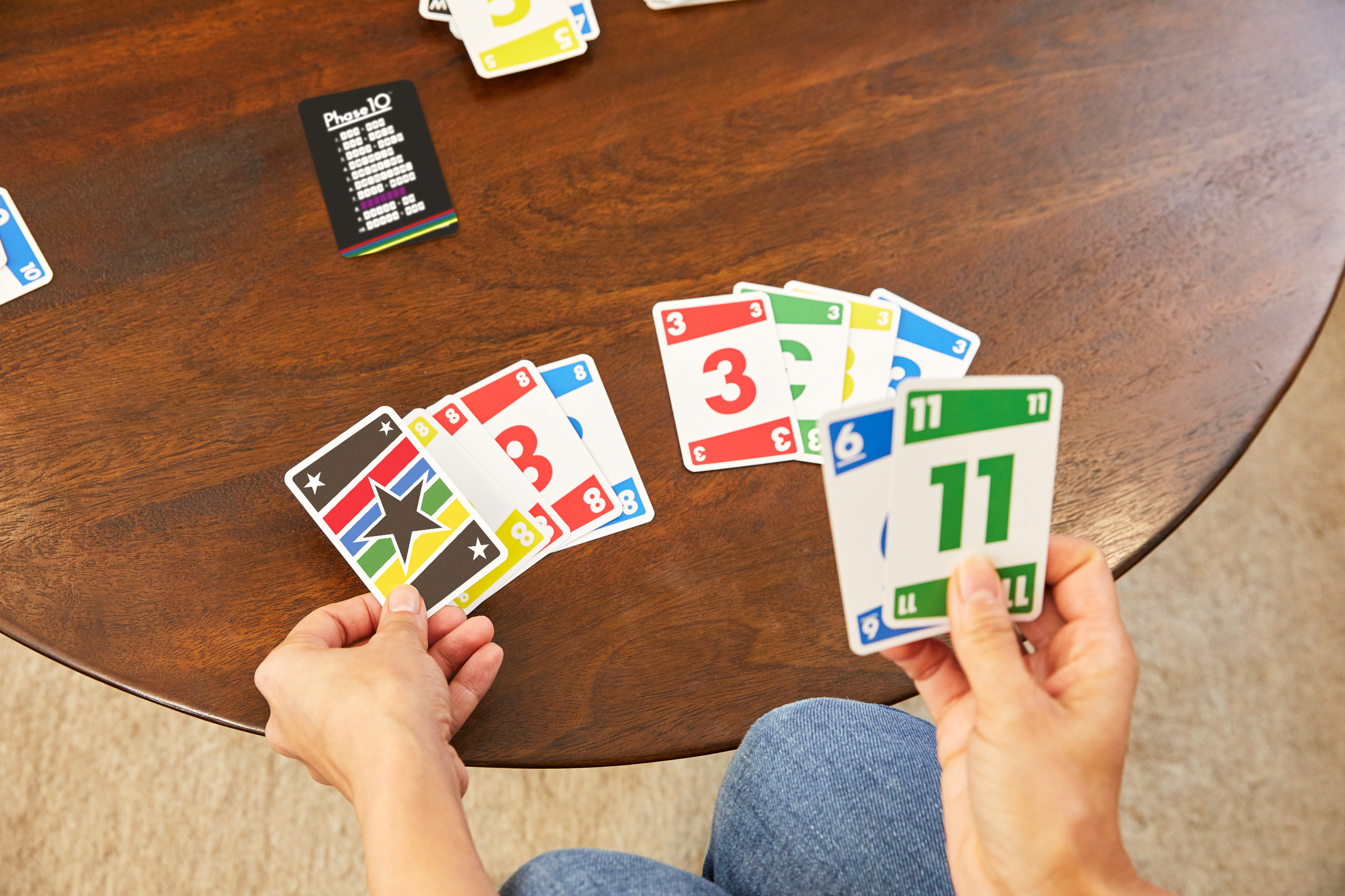Mains tenant des cartes Phase 10 sur une table en bois. Cartes avec chiffres et couleurs visibles.