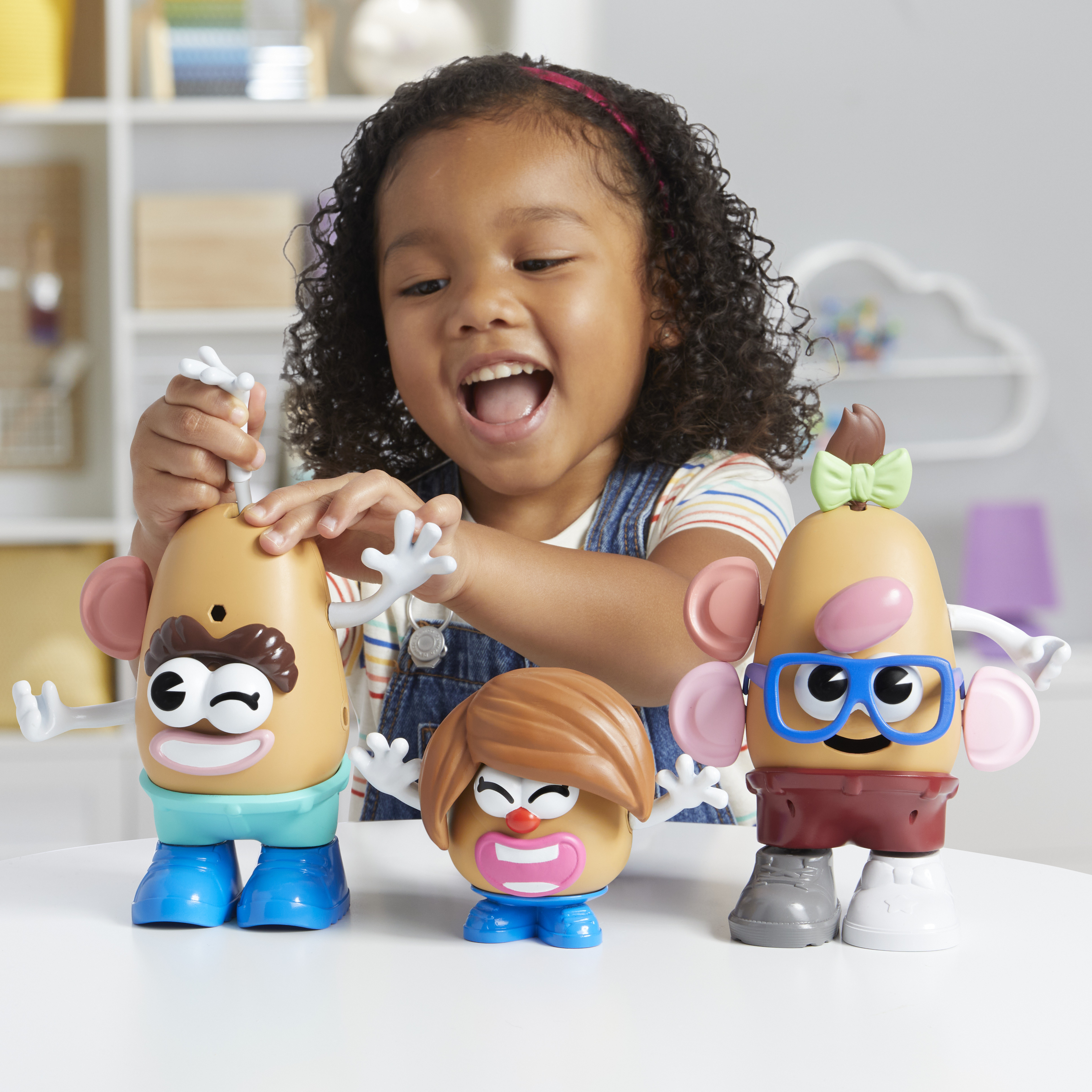 Dziewczynka uśmiecha się do trzech zabawek Mr. Potato Head. Każda ma inną twarz, włosy i akcesoria. Zabawki siedzą na białym stole.