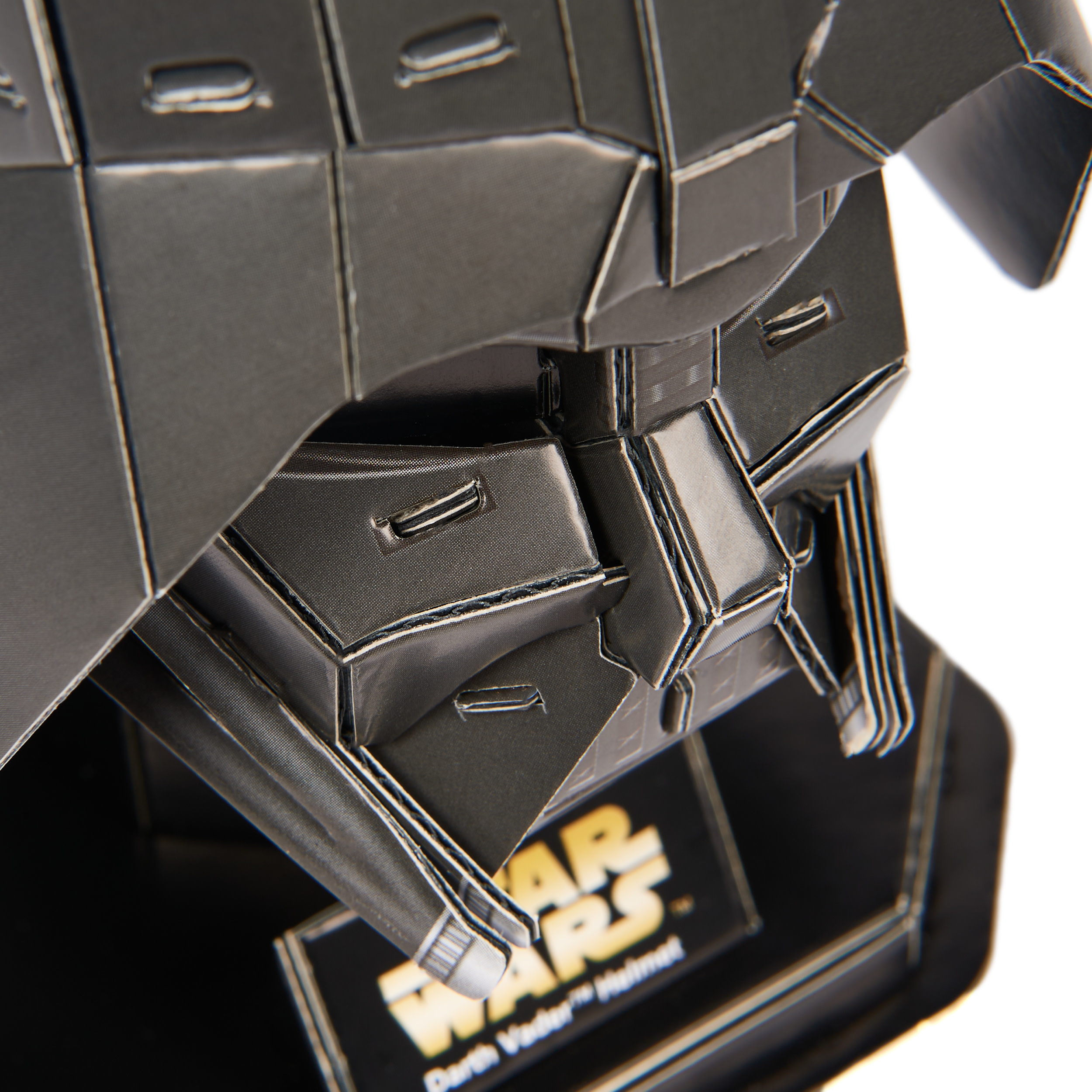 Close-up of a Darth Vader helmet model. It is dark gray with visible seams and rivets. The Star Wars logo is visible.