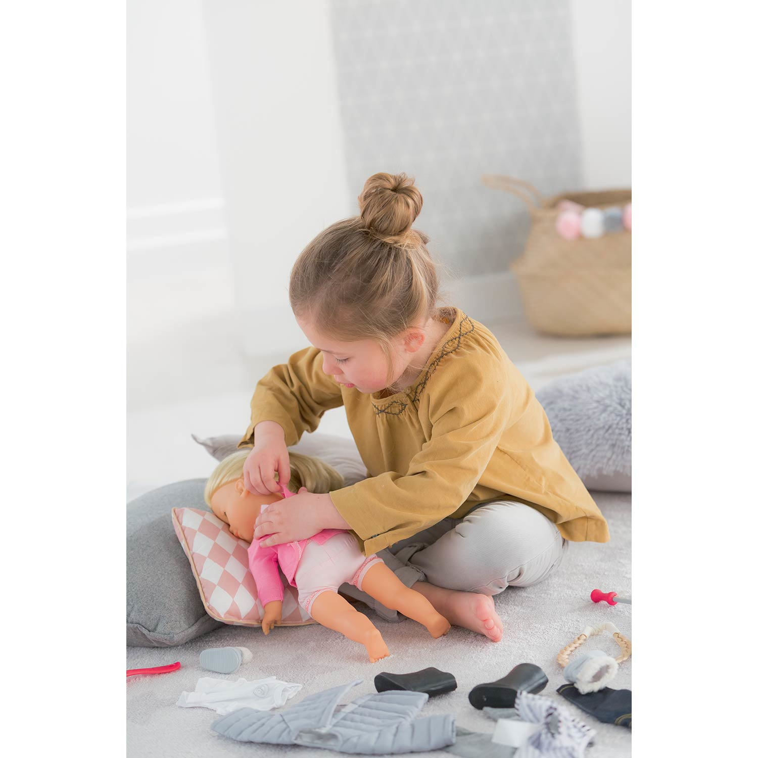 Dziewczynka bawi się lalką. Lalka leży na poduszce, a dziewczynka ją ubiera. Zabawki i ubrania są rozrzucone.