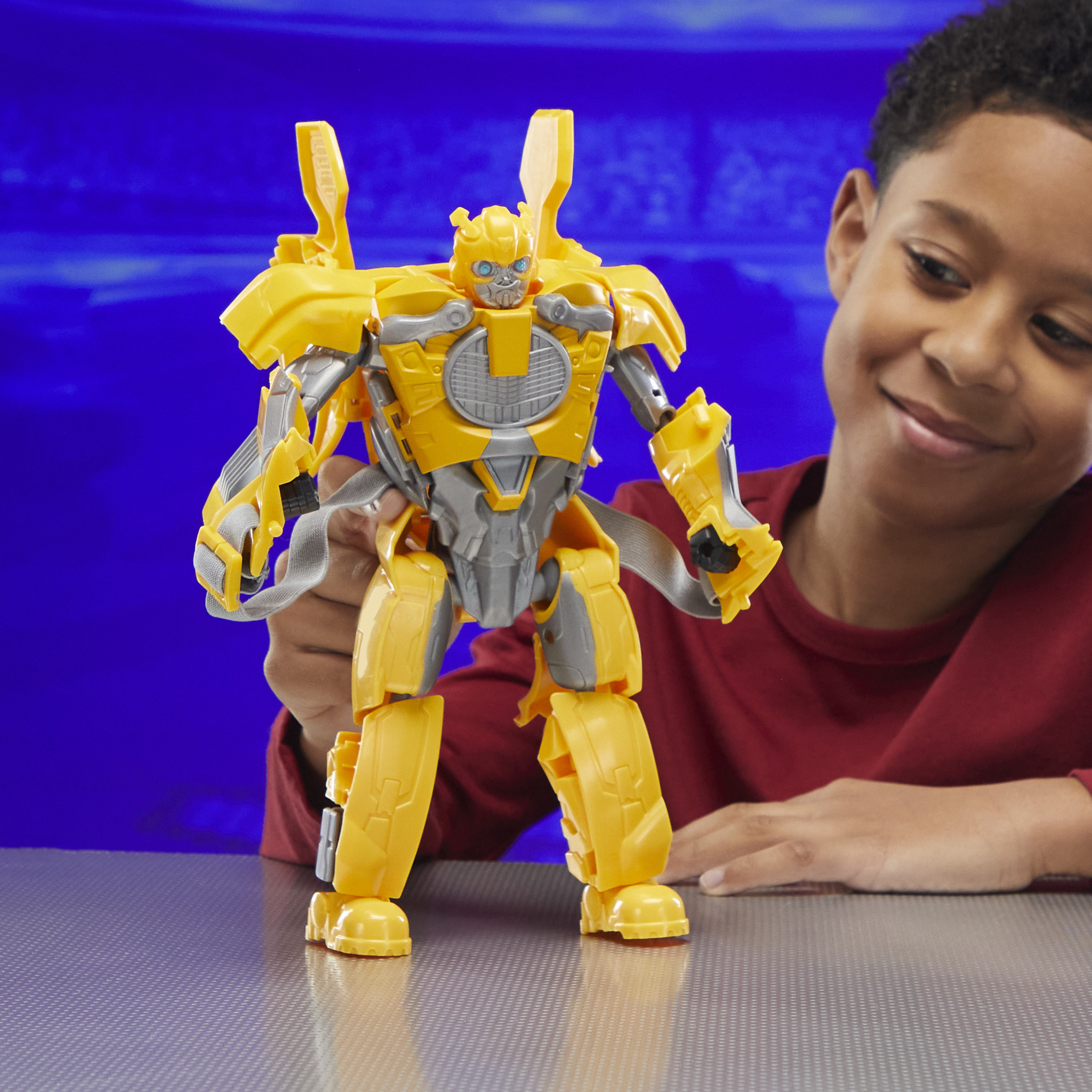 Un niño sostiene un juguete amarillo de Bumblebee. El juguete está sobre una superficie gris contra un fondo azul, y el niño sonríe.