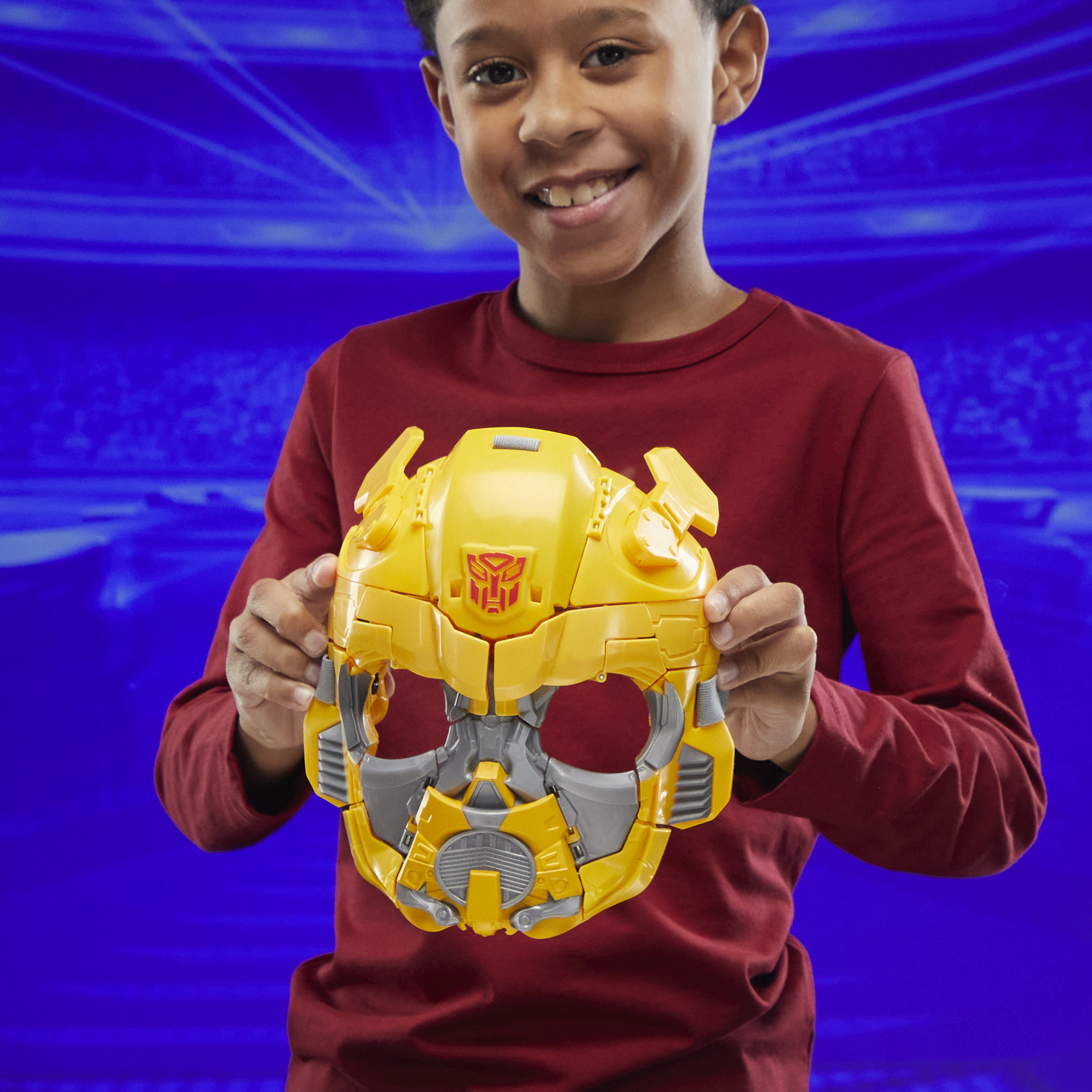 Un niño sostiene una máscara amarilla de Bumblebee. Sonríe. El fondo es azul.