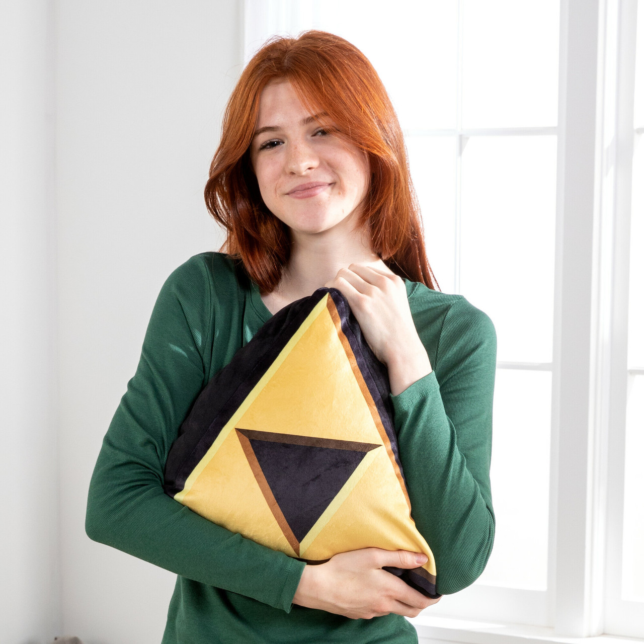 Kobieta z rudymi włosami trzyma poduszkę z żółtym trójkątnym wzorem. Ma na sobie zieloną koszulę i uśmiecha się. W tle widać okno.
