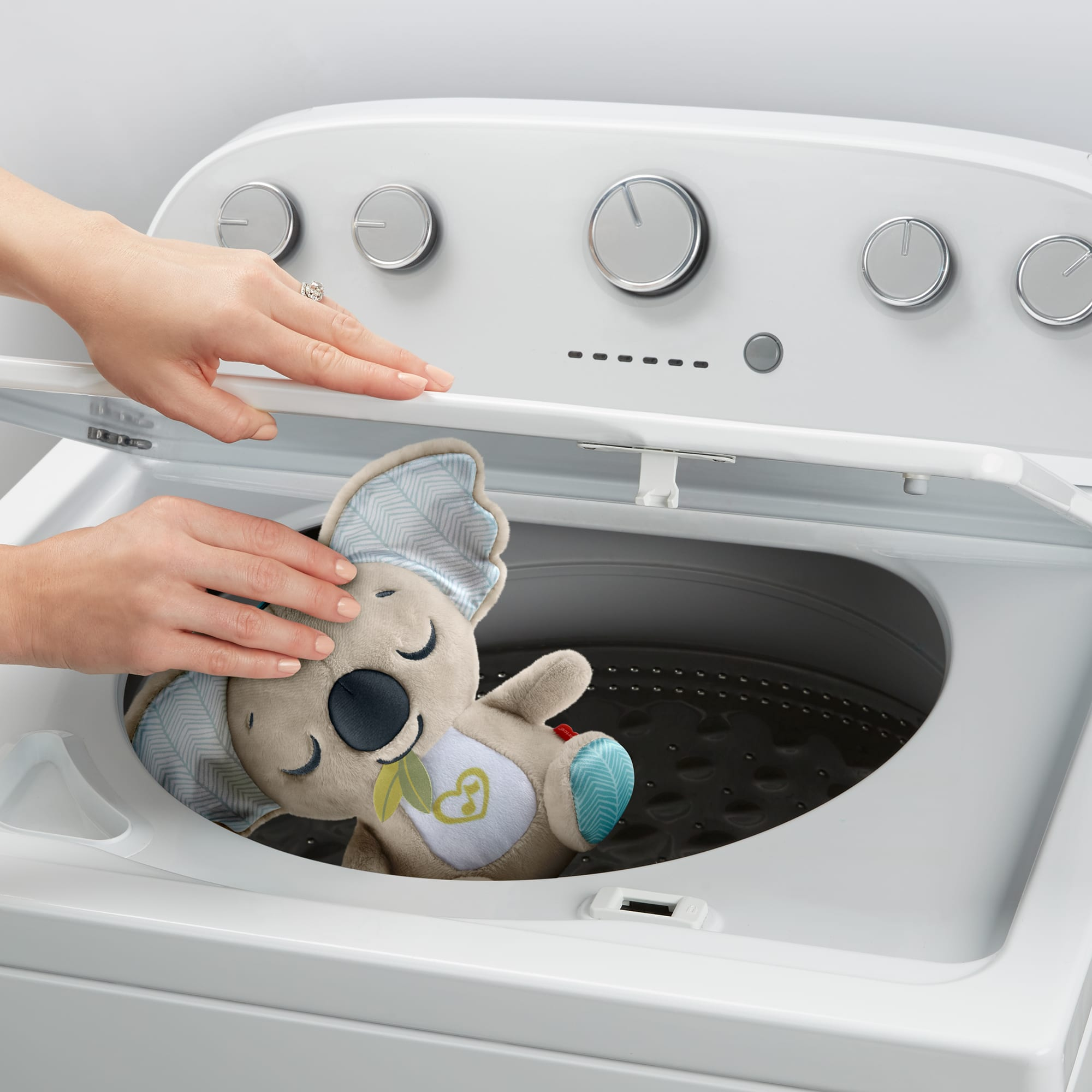 Osoba umieszcza pluszowego misia koala w pralce. Pralka jest biała, a koala szary i niebieski.