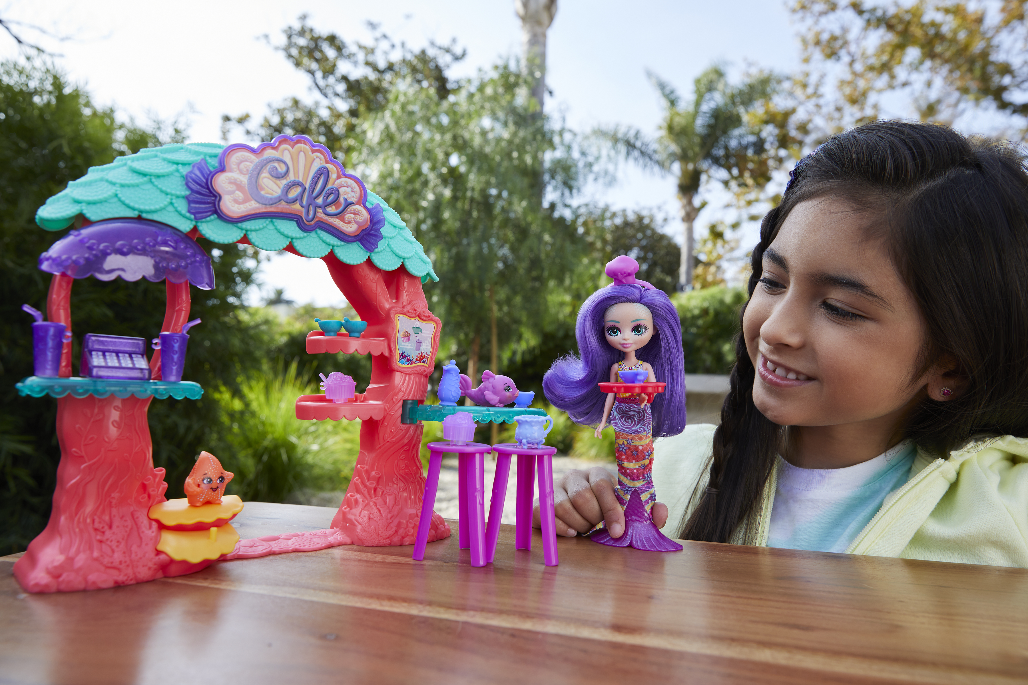 Dziewczyna uśmiecha się do lalki syreny obok zabawkowego kawiarni. W tle drzewa.