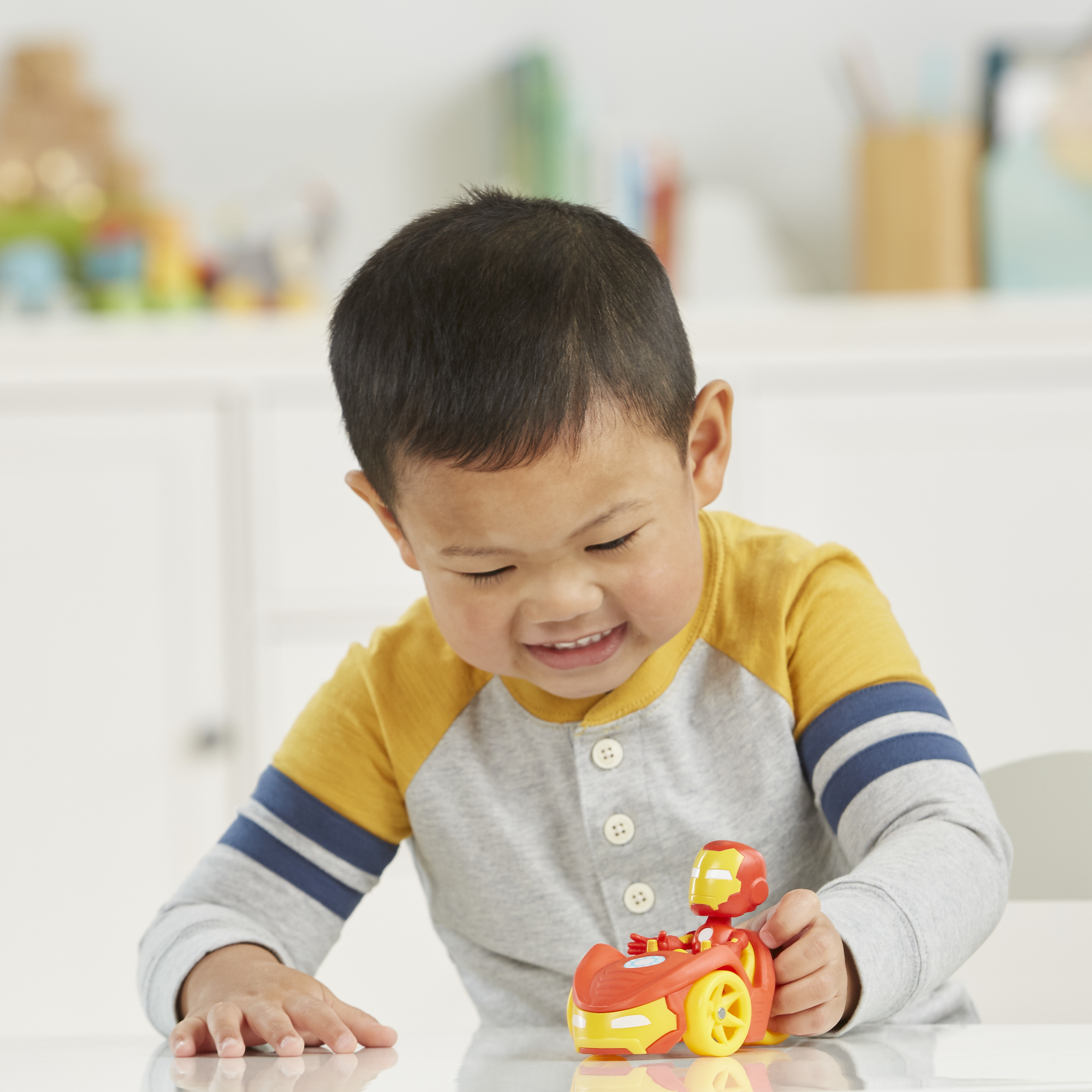 Uśmiechnięty chłopiec patrzy na czerwono-żółty samochodzik. Ma na sobie szarą koszulę z żółtymi rękawami.