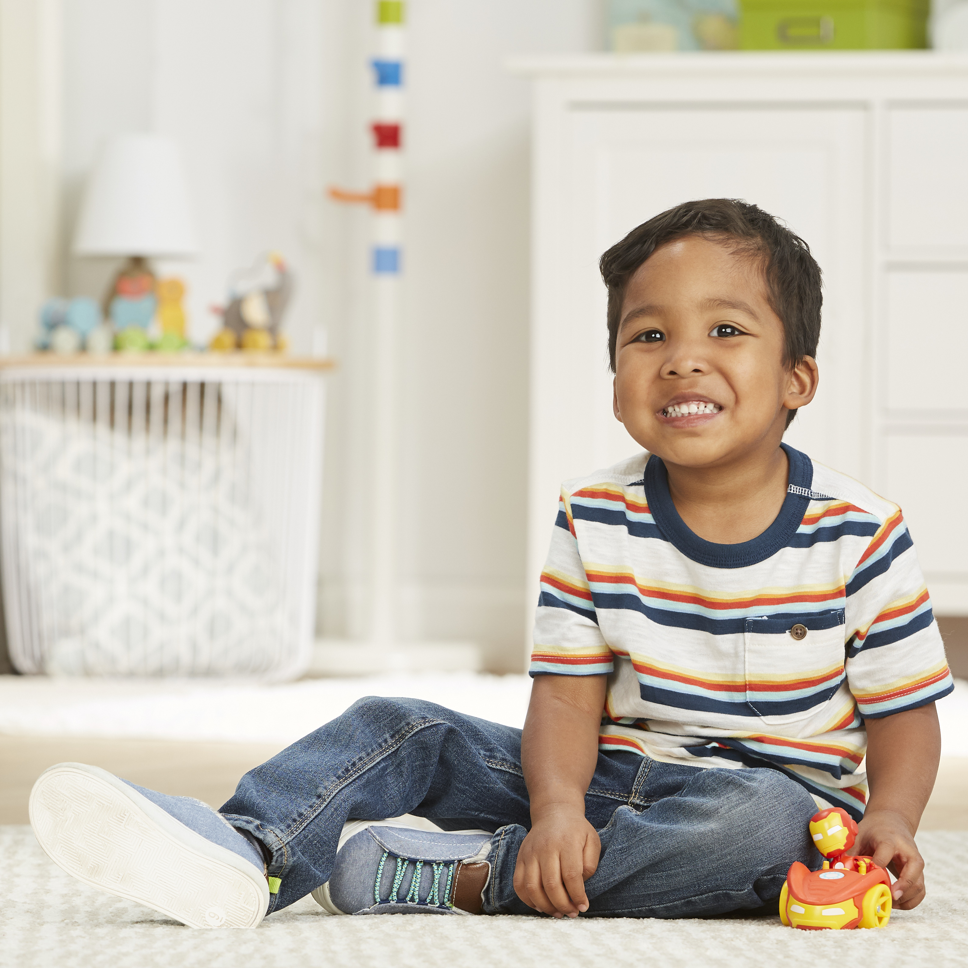 Uśmiechnięty chłopiec w paski siedzi z samochodzikiem. Biała szafka i zabawki w tle.