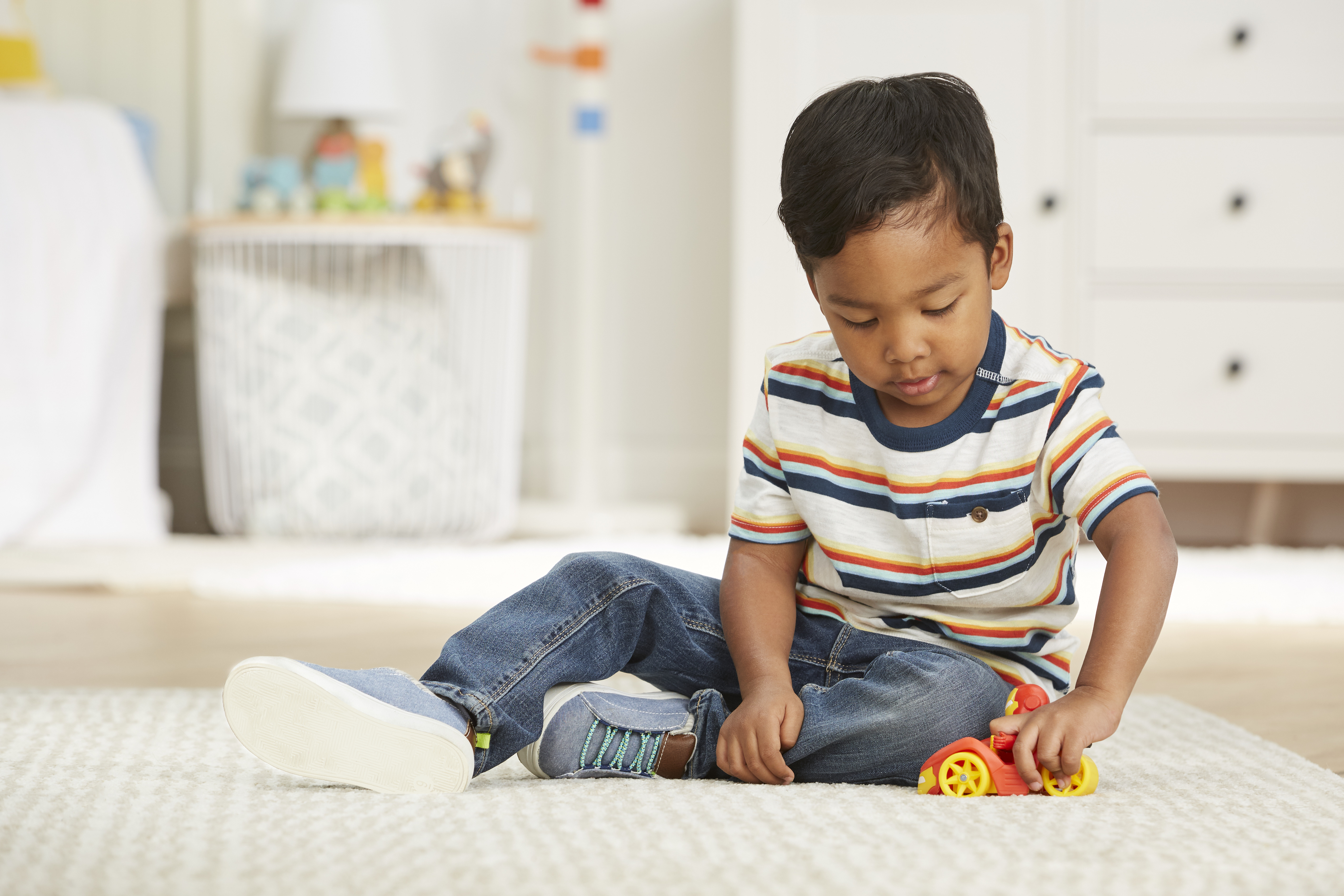 Chłopiec siedzi na dywanie, bawiąc się zabawką-samochodem. Ma na sobie dżinsy i koszulę w paski. W tle biała komoda.