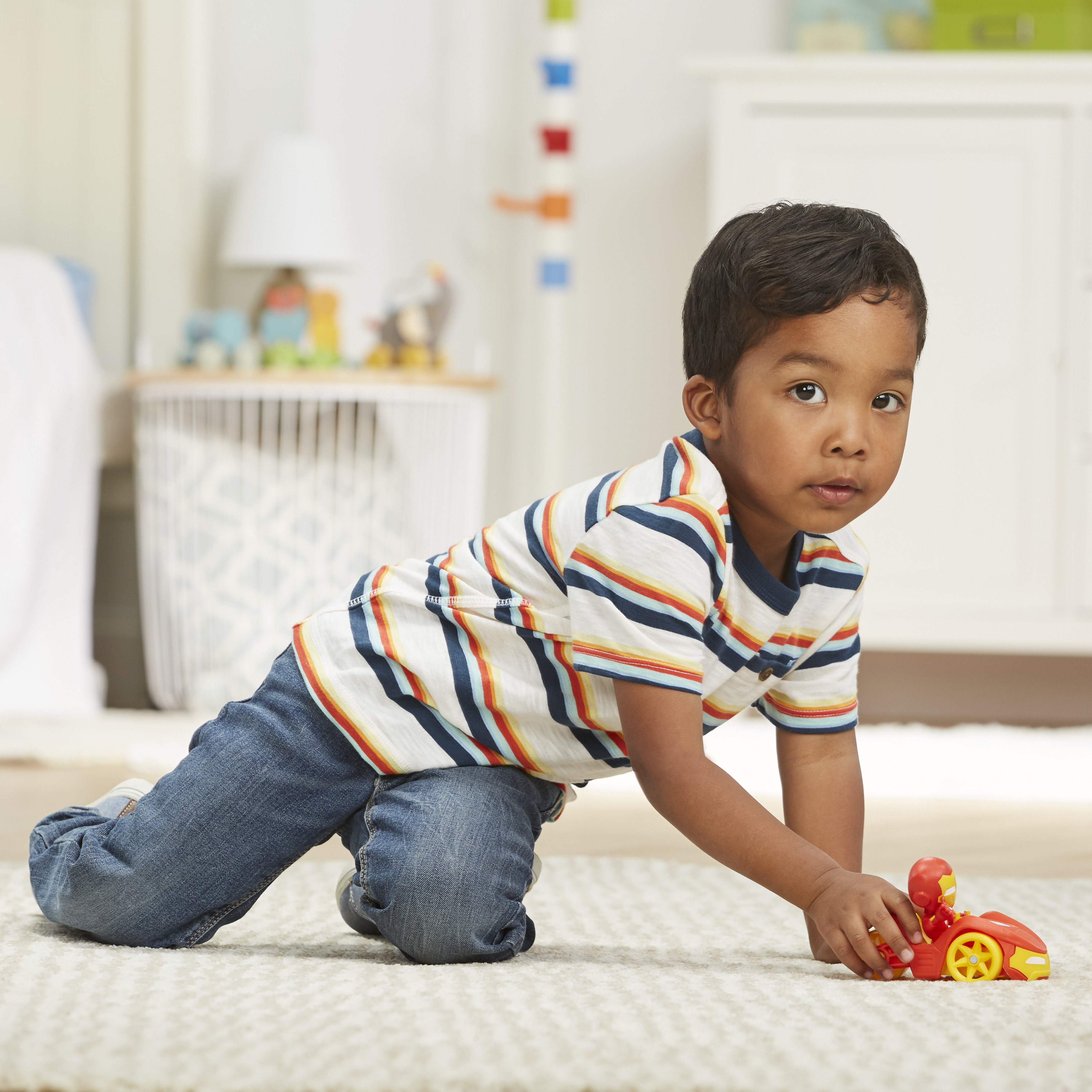 Chłopiec w paski bawi się czerwoną zabawką. Jest na dywanie w pokoju.