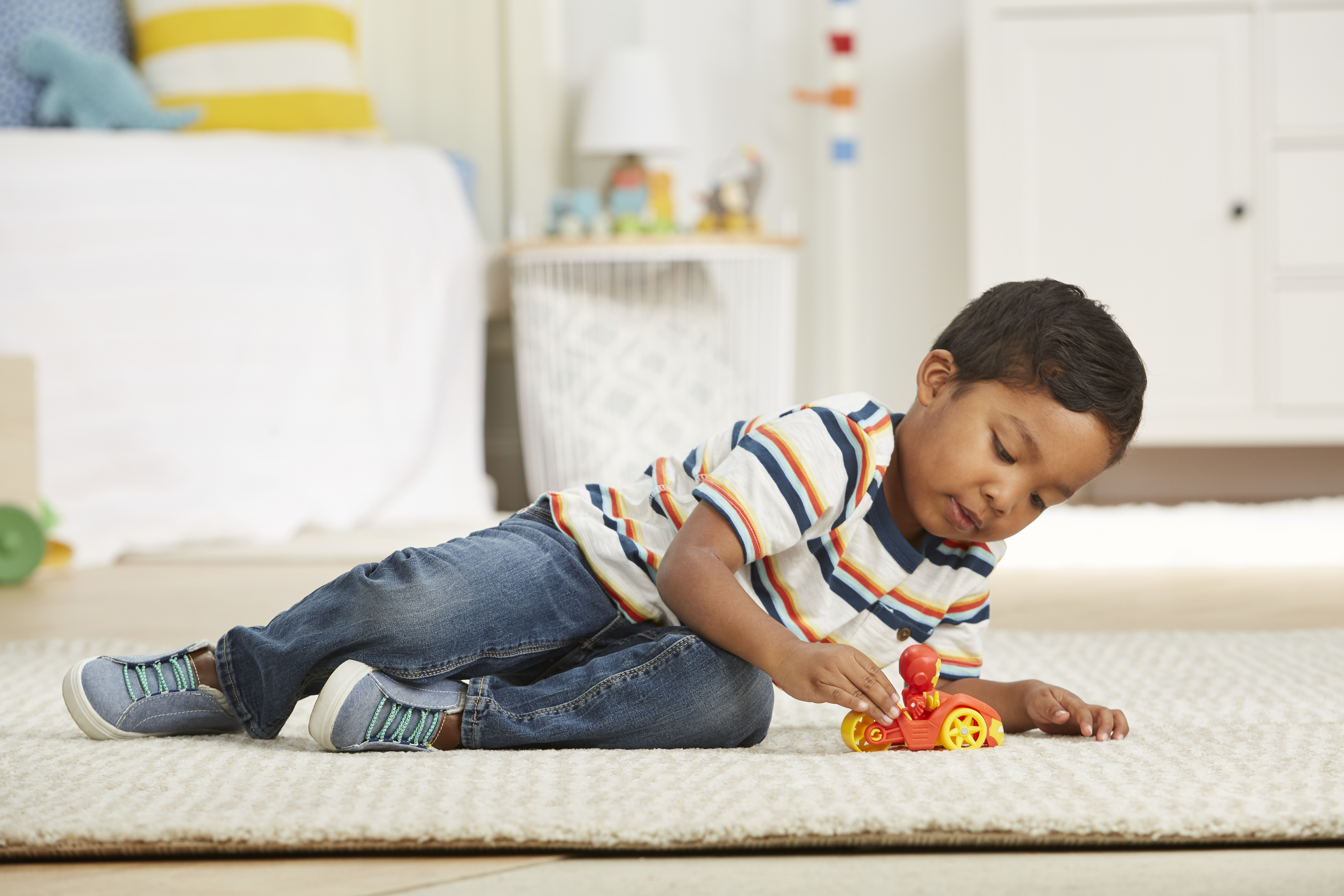 Mały chłopiec bawi się czerwoną i żółtą zabawką-samochodem na dywanie. Ma na sobie dżinsy i koszulę w paski.