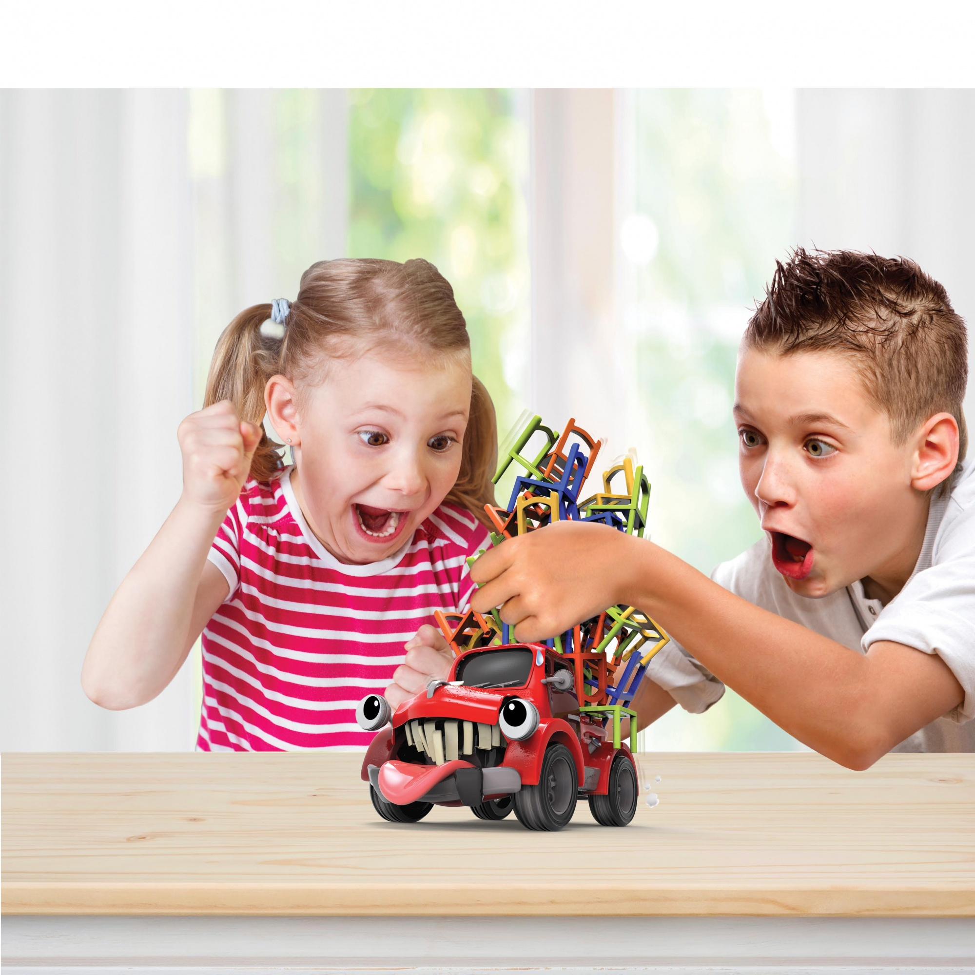 Dwoje dzieci jest zaskoczonych przez zabawkową ciężarówkę przewożącą kolorowe krzesła. Ciężarówka ma twarz. Tło to okno.