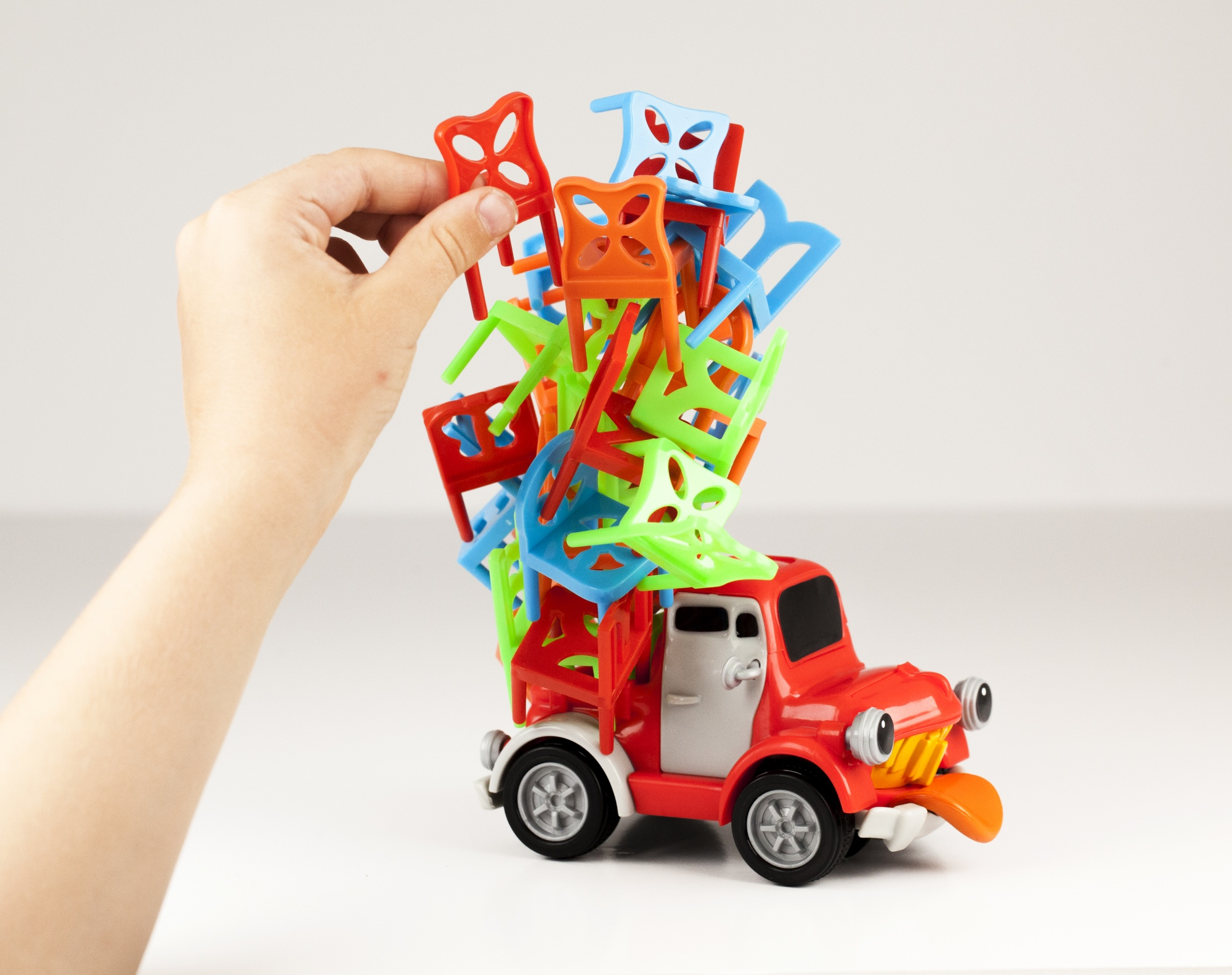 Ręka umieszcza czerwone krzesło na ciężarówce przewożącej kolorowe krzesła. Ciężarówka jest czerwona z twarzą.