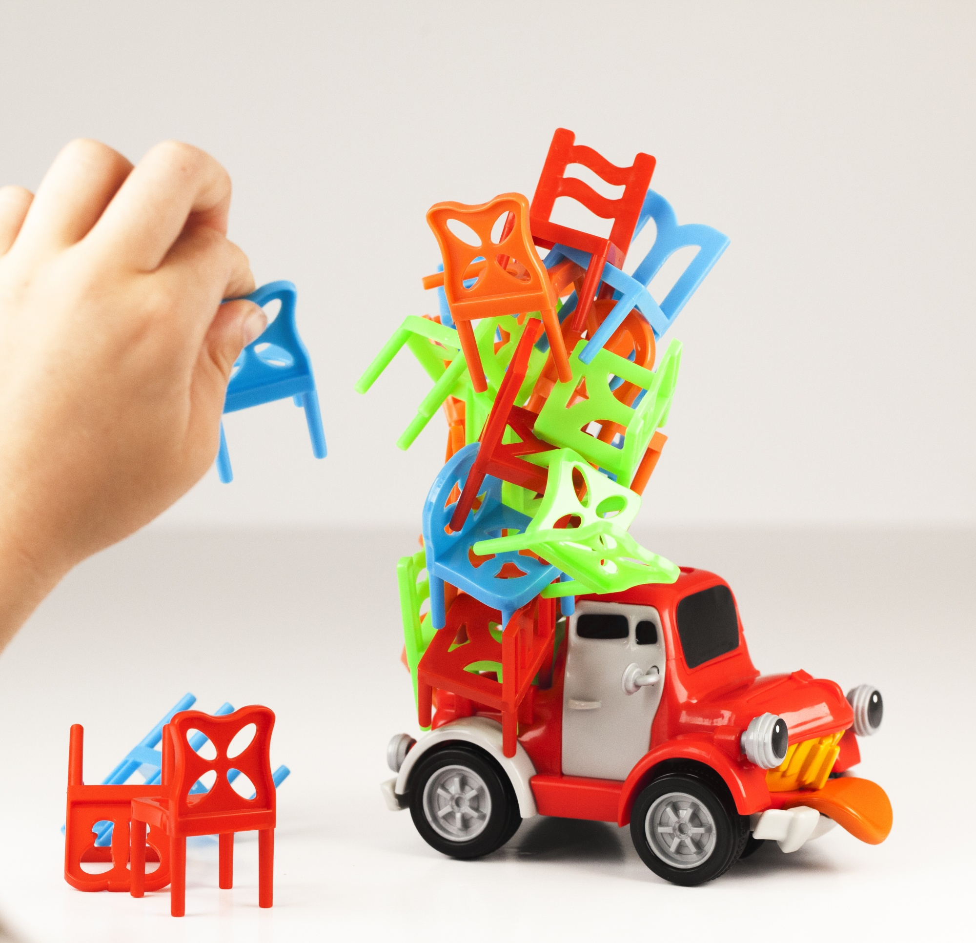 Ręka umieszcza niebieskie krzesło na stosie kolorowych krzeseł na czerwonej zabawce ciężarówki. Krzesła są zielone, pomarańczowe i czerwone.