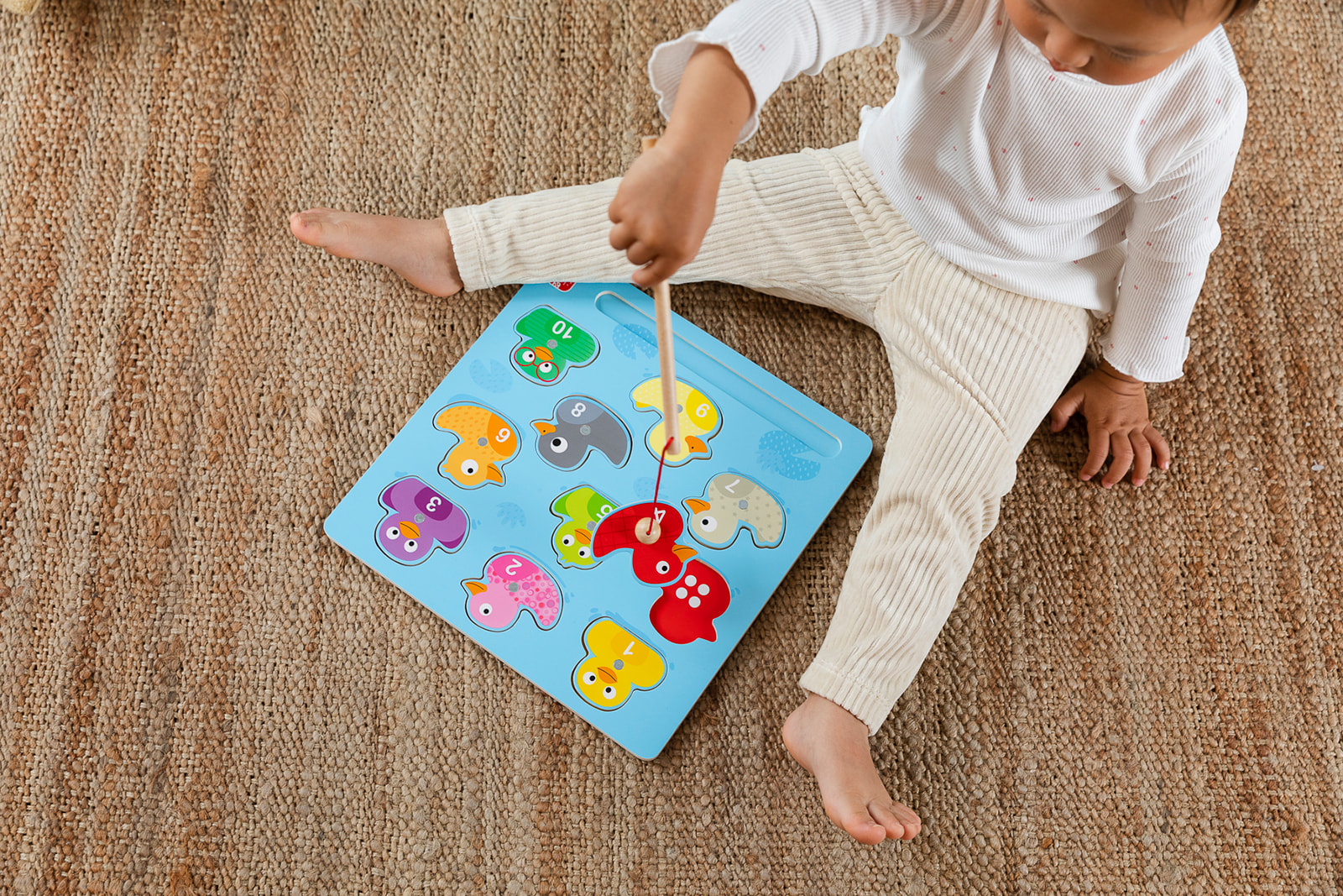 Dziecko bawi się niebieską układanką z kolorowymi kształtami ptaków na tkanym dywanie. Dziecko ma na sobie białą górę i beżowe spodnie.